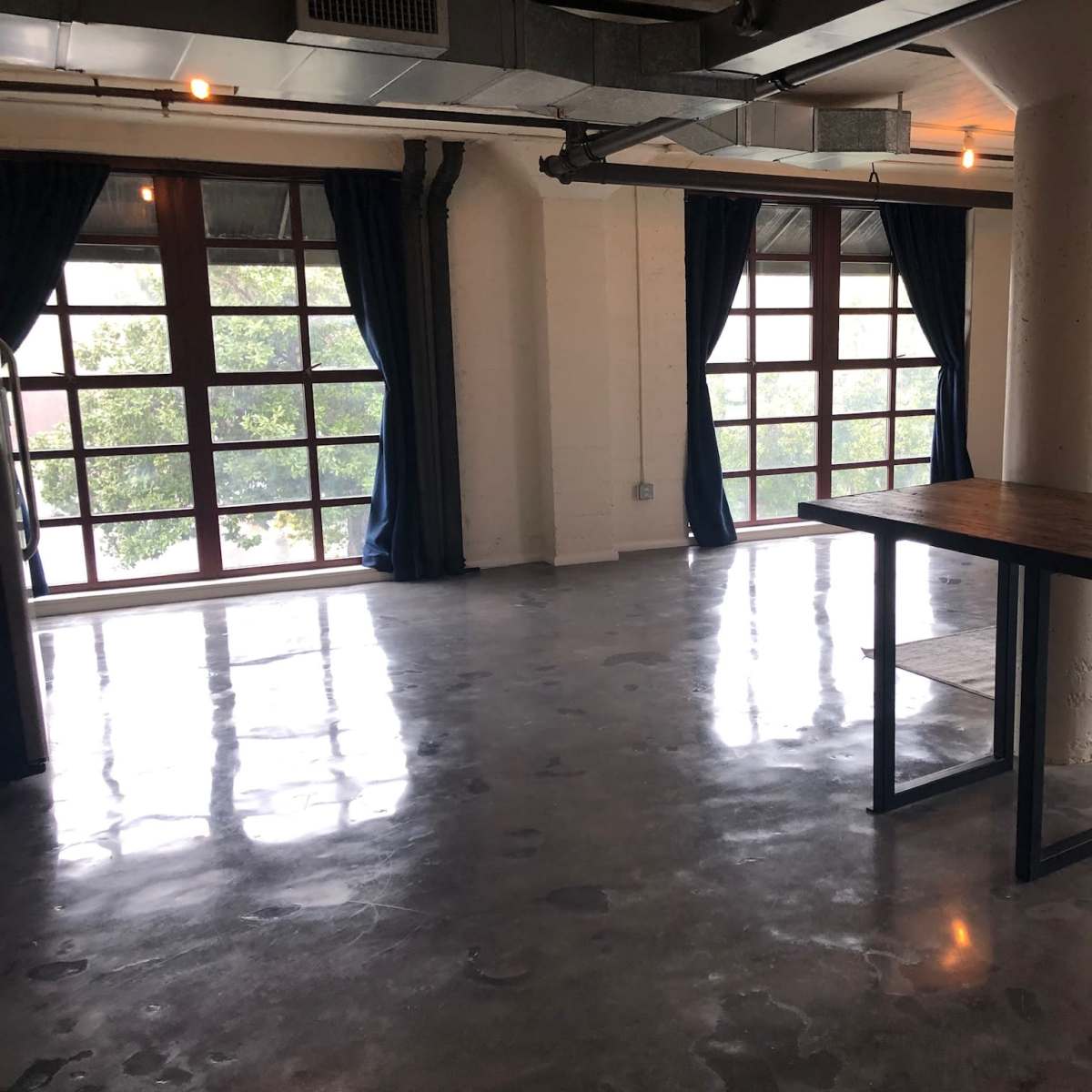 A spacious, empty room with polished concrete floors, large windows, and a wooden table in the center.