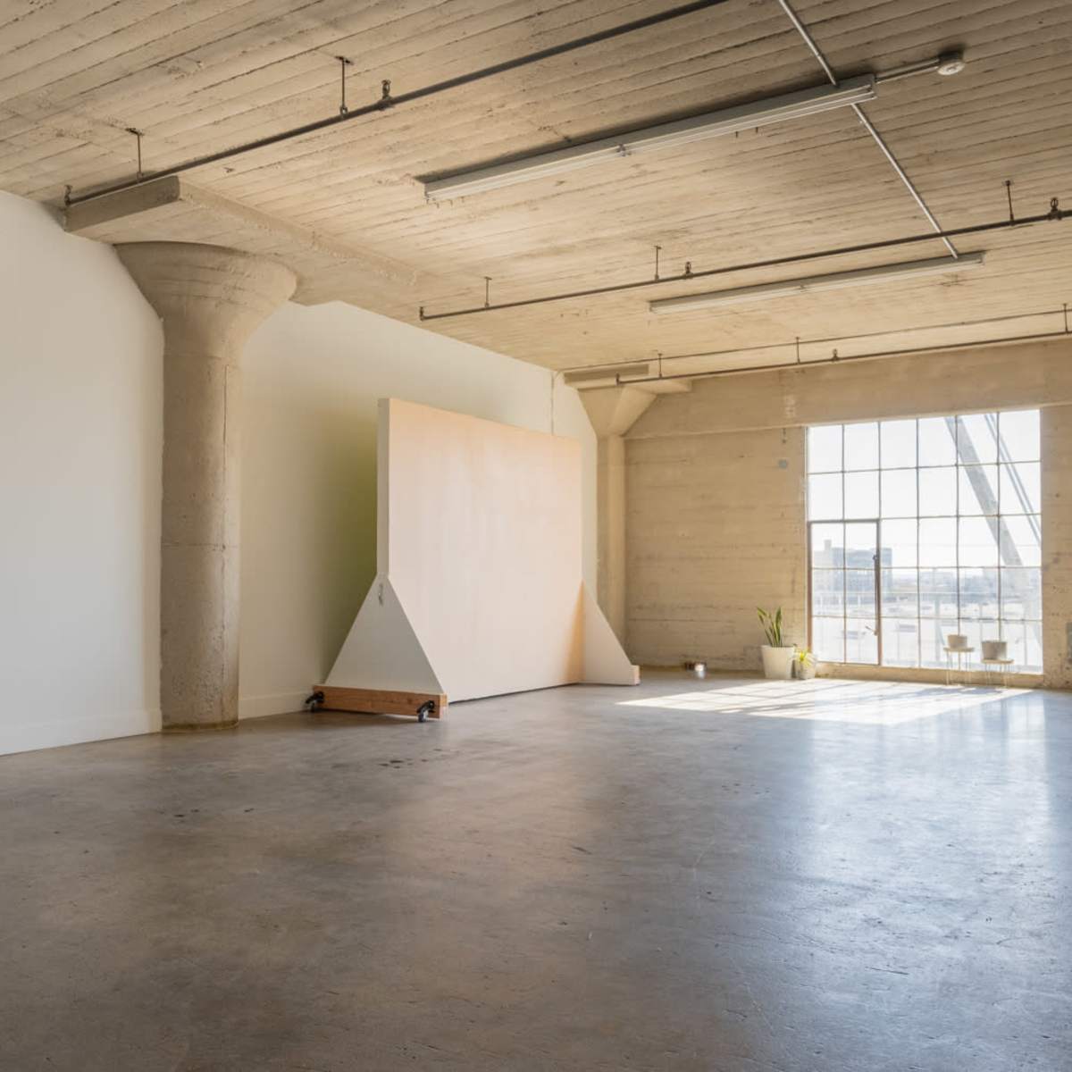 A spacious, empty room with concrete walls, high ceilings, and a large window letting in natural light, featuring a large, triangular wooden panel against one wall.