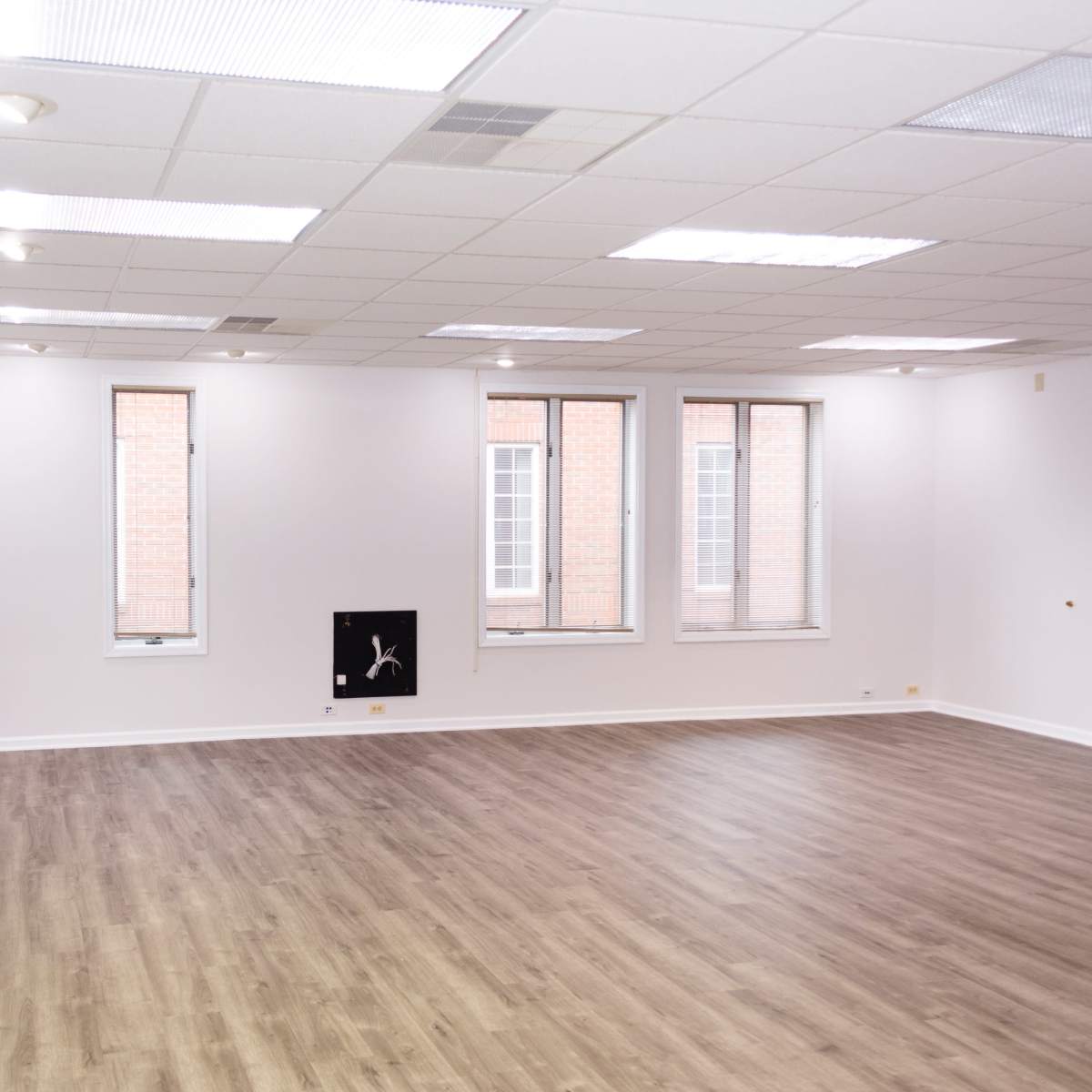An empty room with wooden flooring and three windows, featuring plain white walls and a small wall-mounted object.