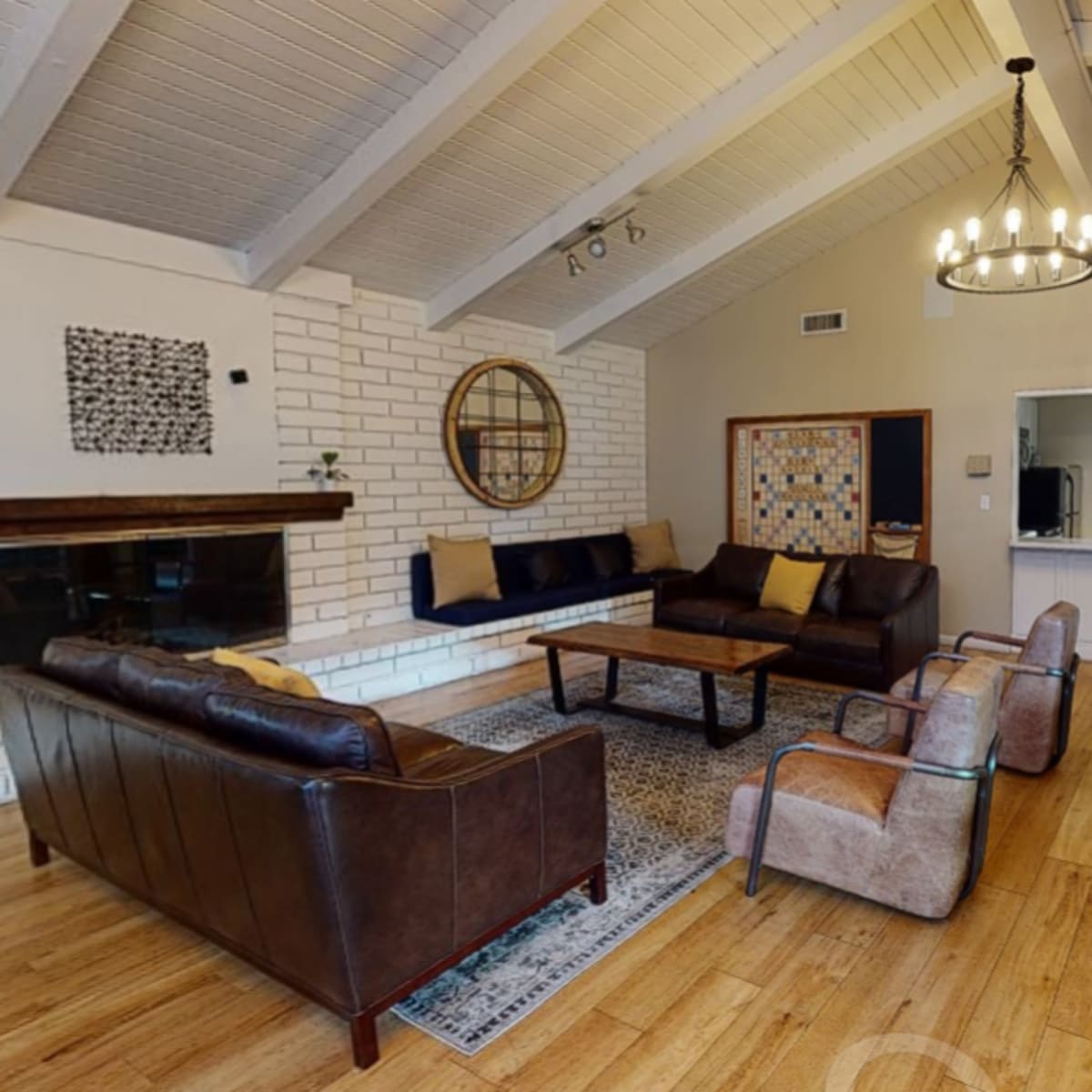 A spacious living room featuring a mix of brown leather sofas, a wooden coffee table, and a stone fireplace, all set against a backdrop of white walls and wooden beams.