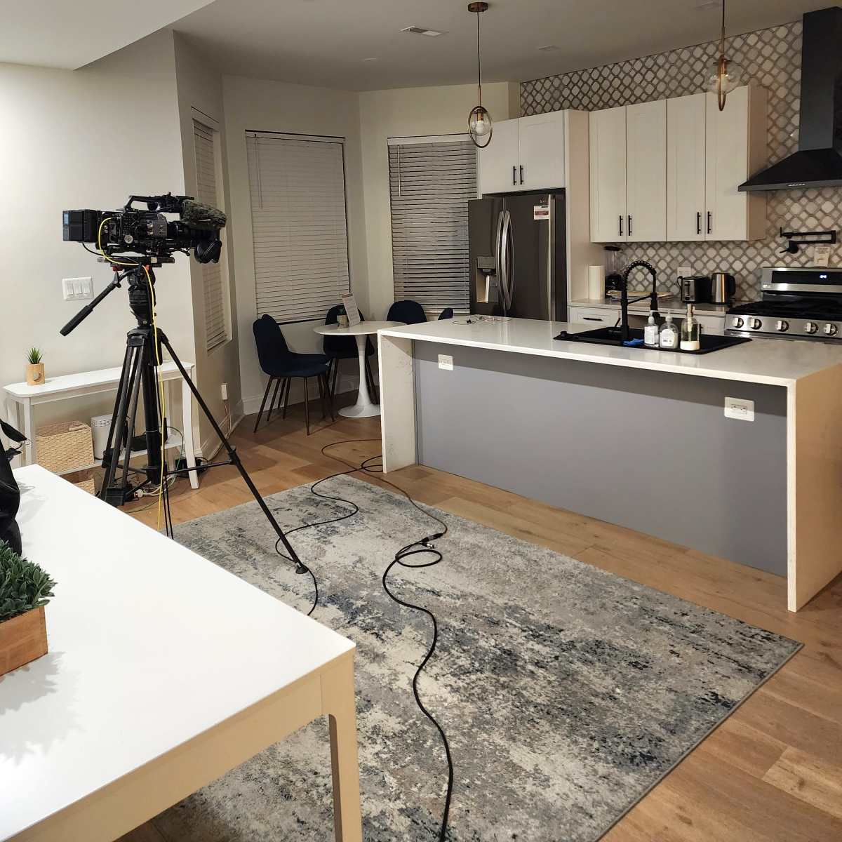 A kitchen is set up for filming, featuring a camera on a tripod, a dining table with chairs, and modern appliances.