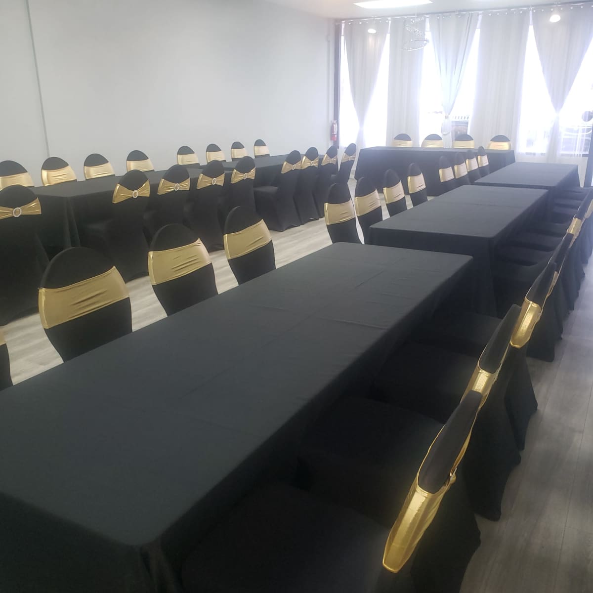 A spacious banquet room features long tables covered with black tablecloths and chairs adorned with gold sashes.