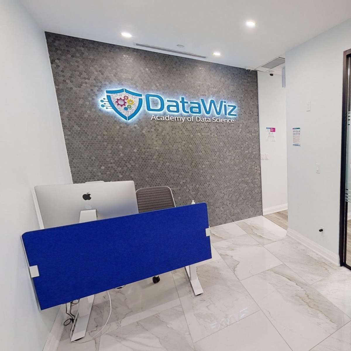 A modern office reception area with the "DataWiz Academy of Data Science" logo on the wall and colorful seating options visible in the foreground.