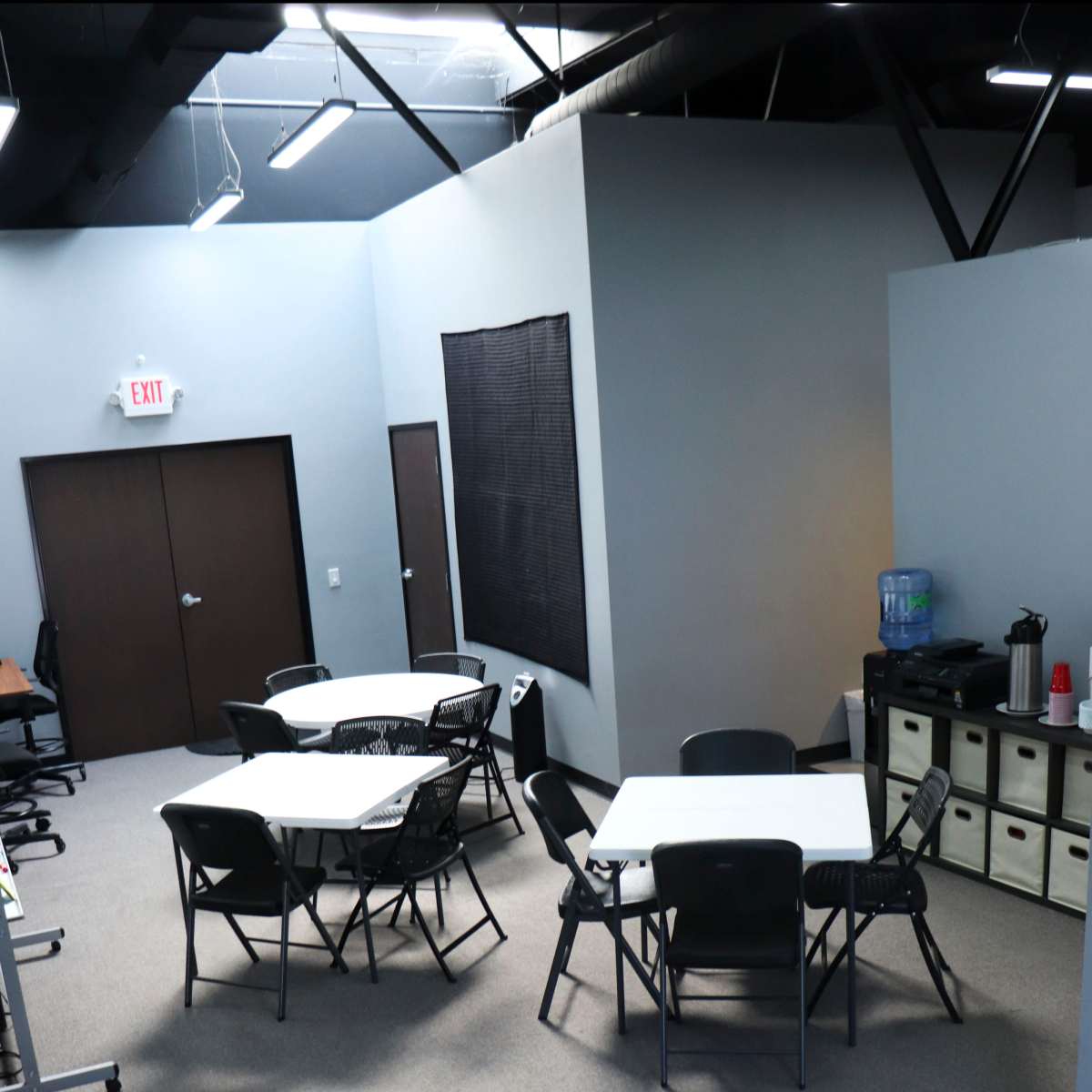 A meeting space with several tables and chairs arranged in a room featuring gray walls and a black window covering.