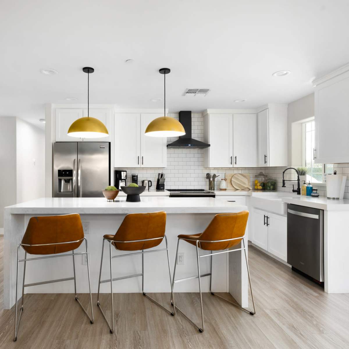 A modern kitchen features white cabinetry, a large island with three bar stools, and two pendant lights above, with a living area visible in the background.