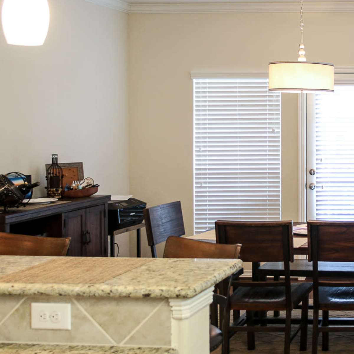 A dining area with a wooden table and chairs, a light fixture overhead, and windows with closed blinds.