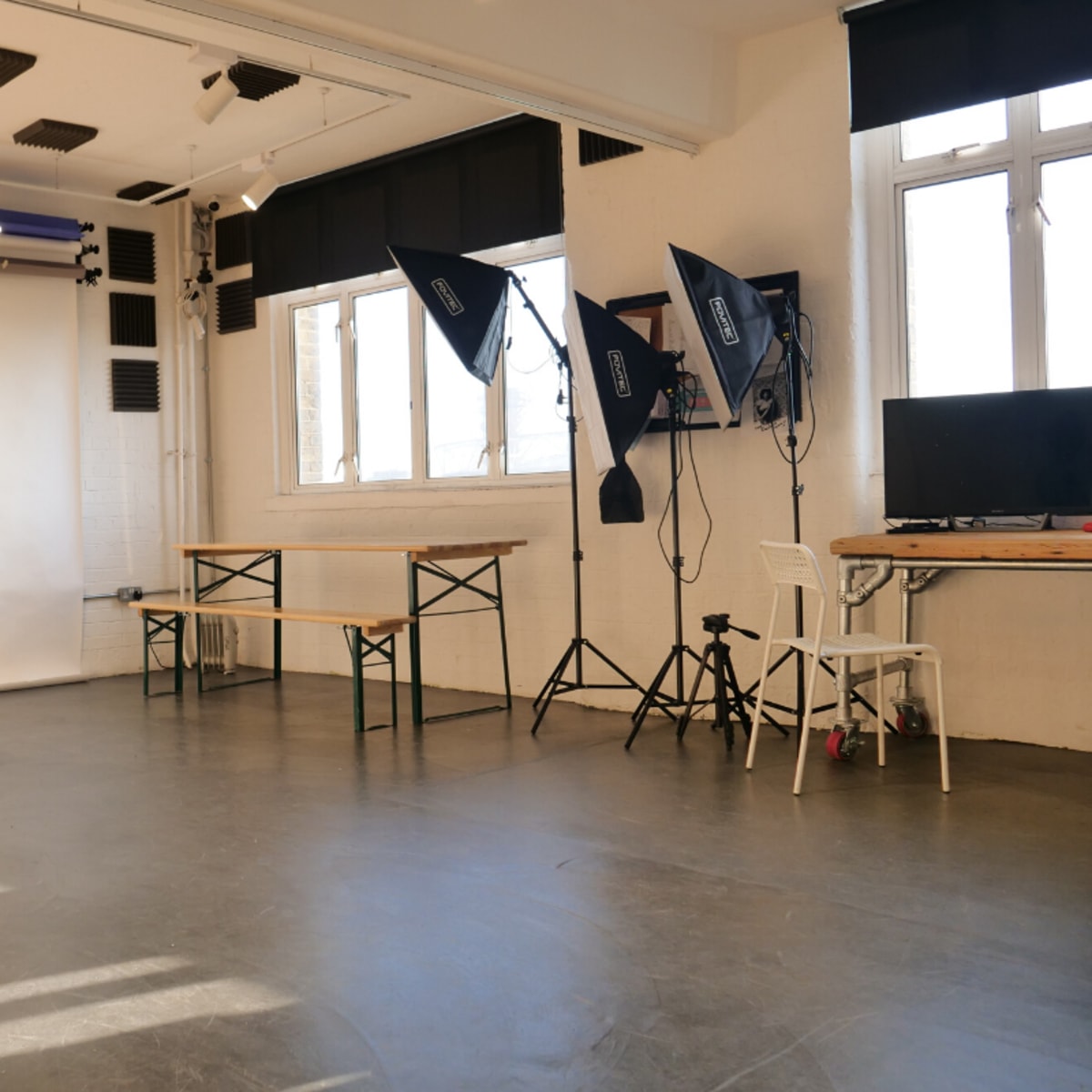 A spacious studio with wooden tables, lighting equipment, and a blank backdrop near large windows.