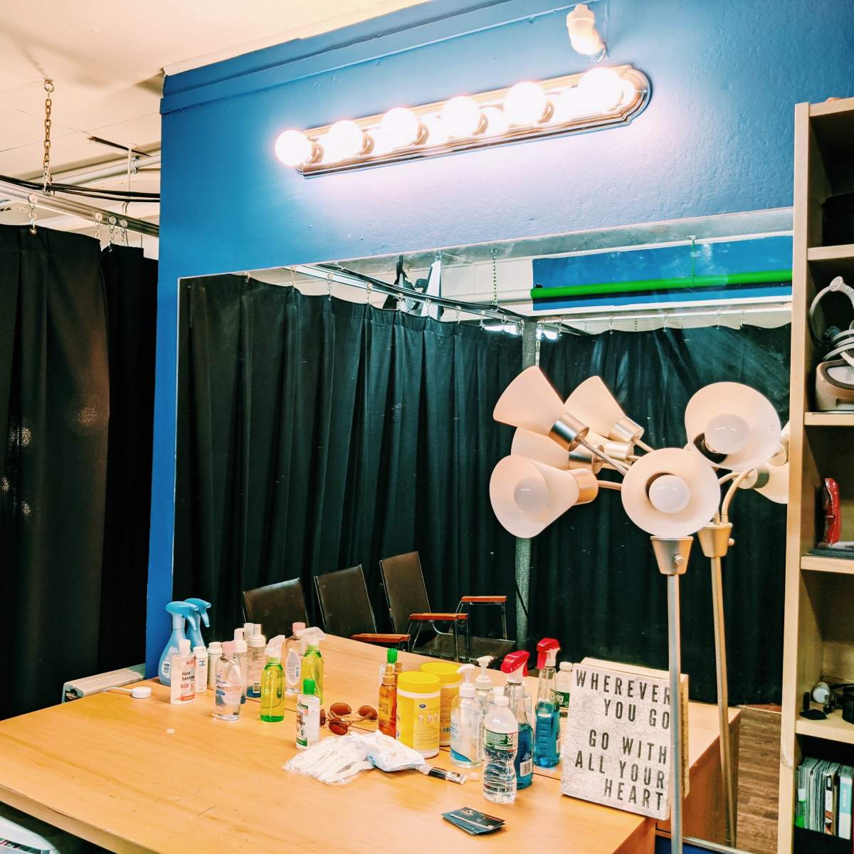 A mirror with a bright light fixture above it, surrounded by a black curtain backdrop and a lamp on a wooden table filled with various bottles and products.