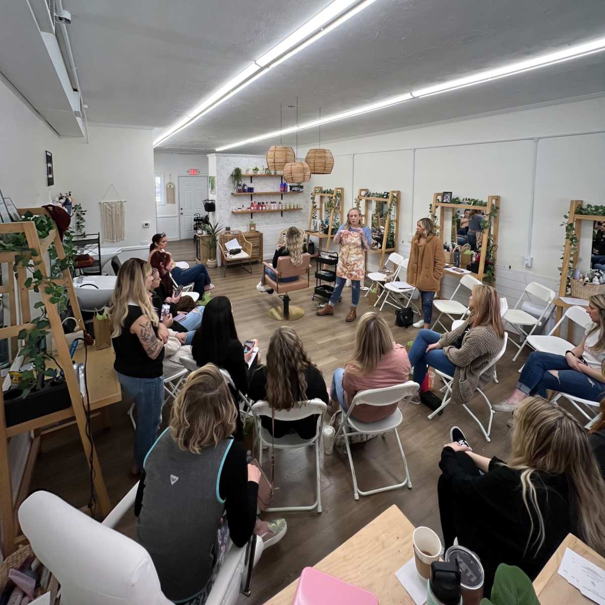 A group of people is seated in a well-lit room with mirrors and plants, while one person stands and speaks to them.