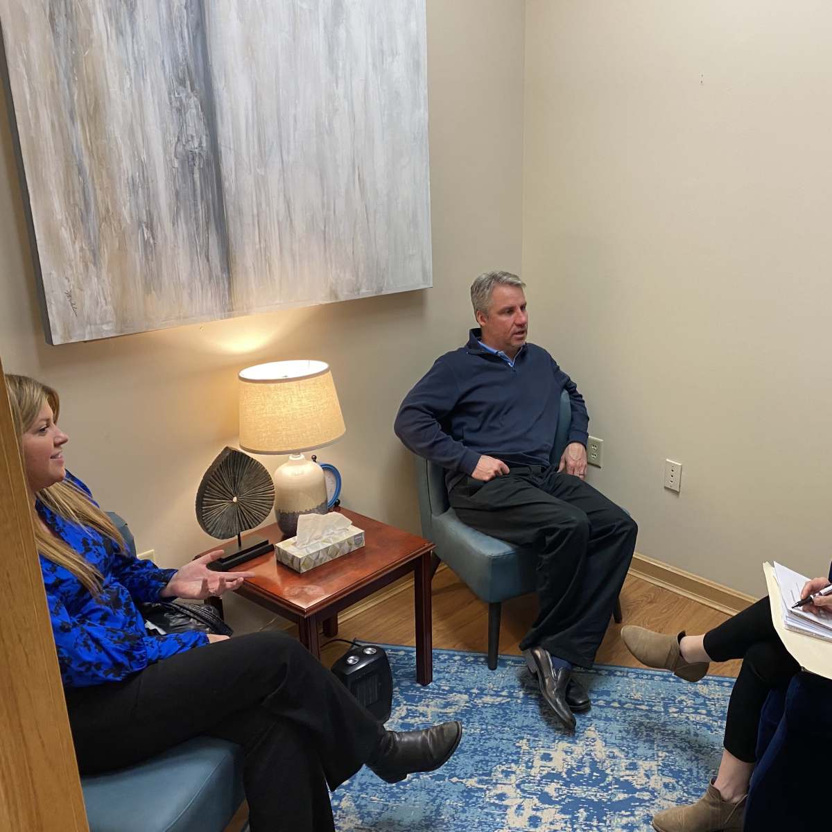 Three people sit in a small, well-lit room, engaging in a discussion, with a painting and a lamp on a side table nearby.