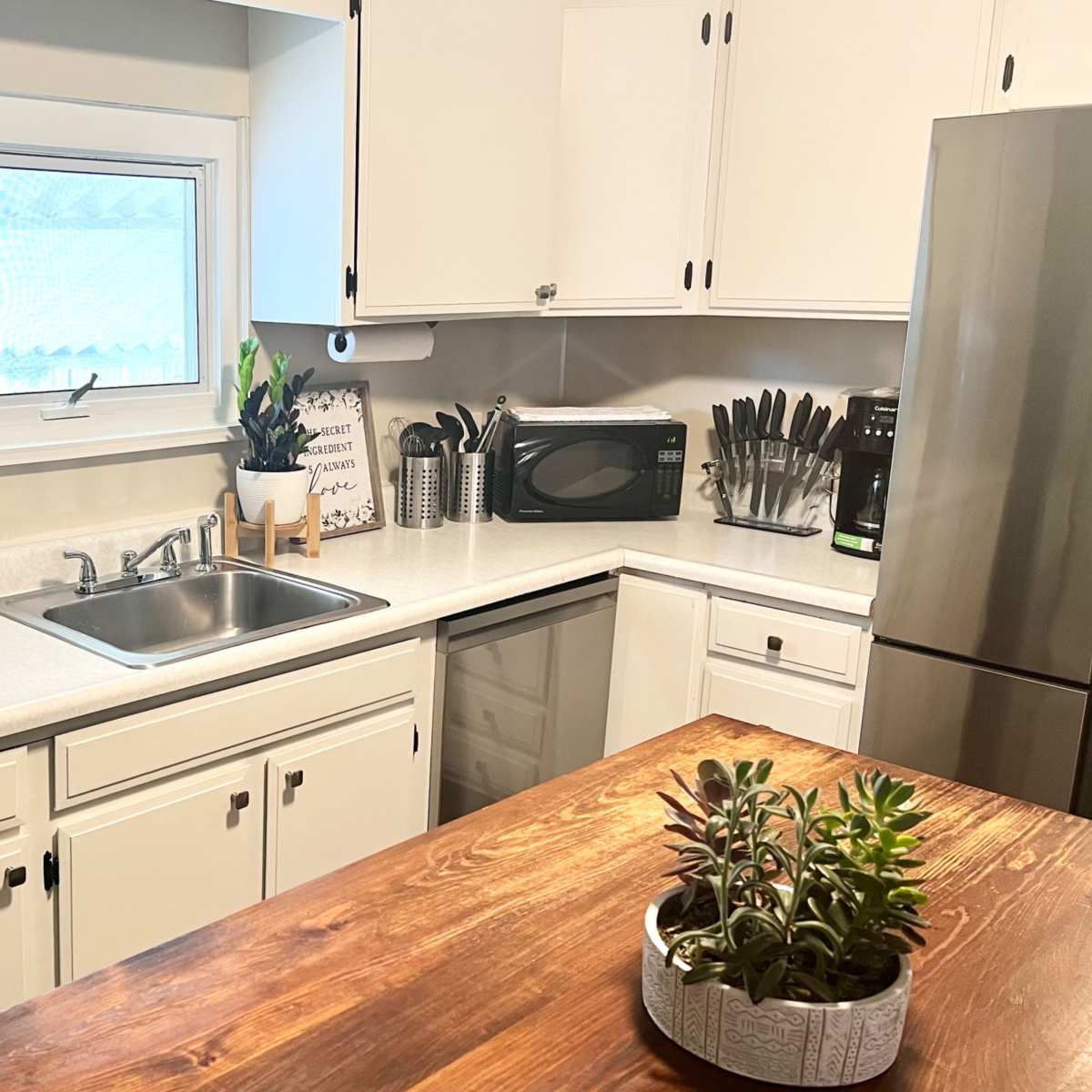 A bright kitchen features white cabinets, a stainless steel refrigerator, a sink, and a wooden island with potted succulents.