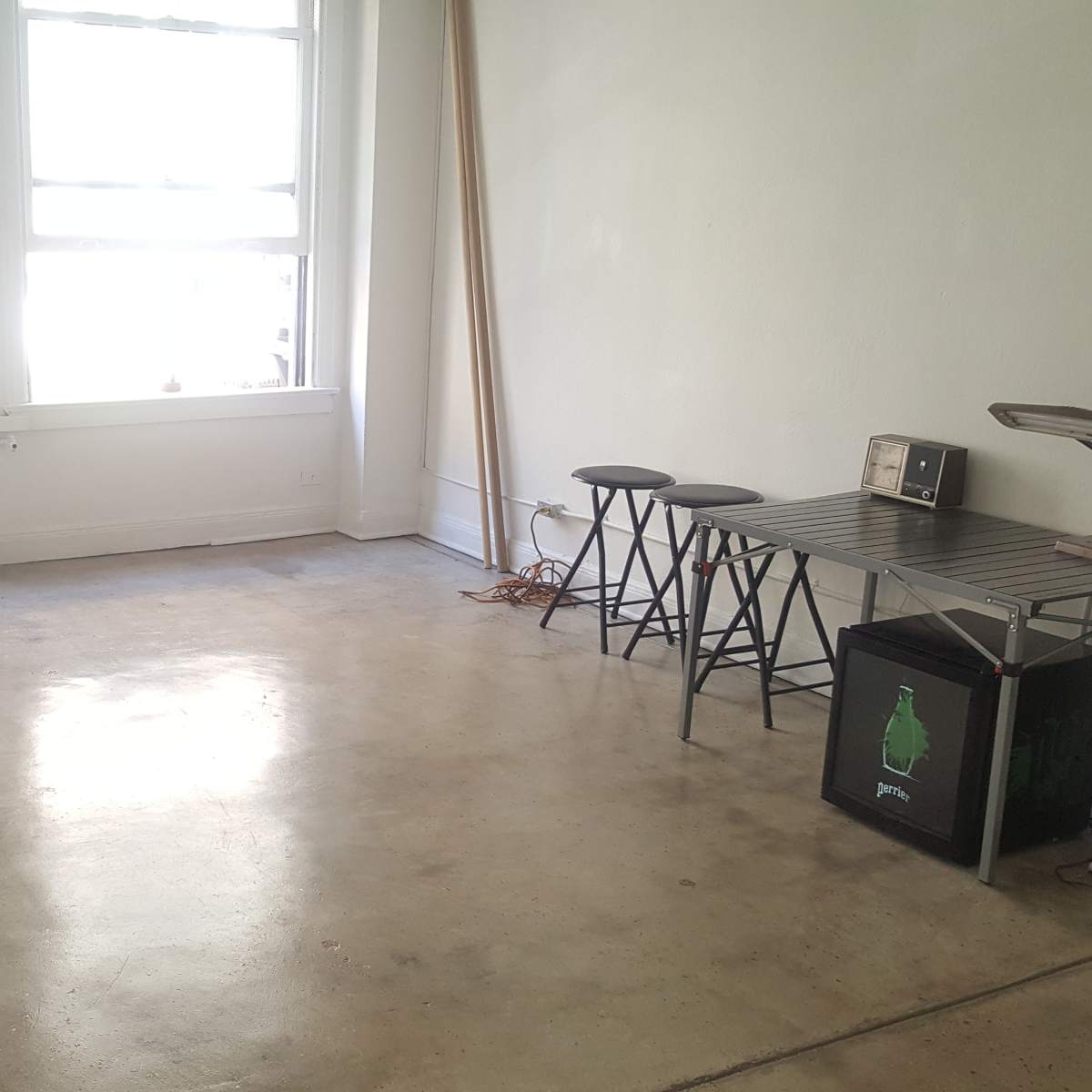 A minimalist room with a concrete floor, two large windows, a small kitchen table, three stools, a microwave, and a compact fridge.