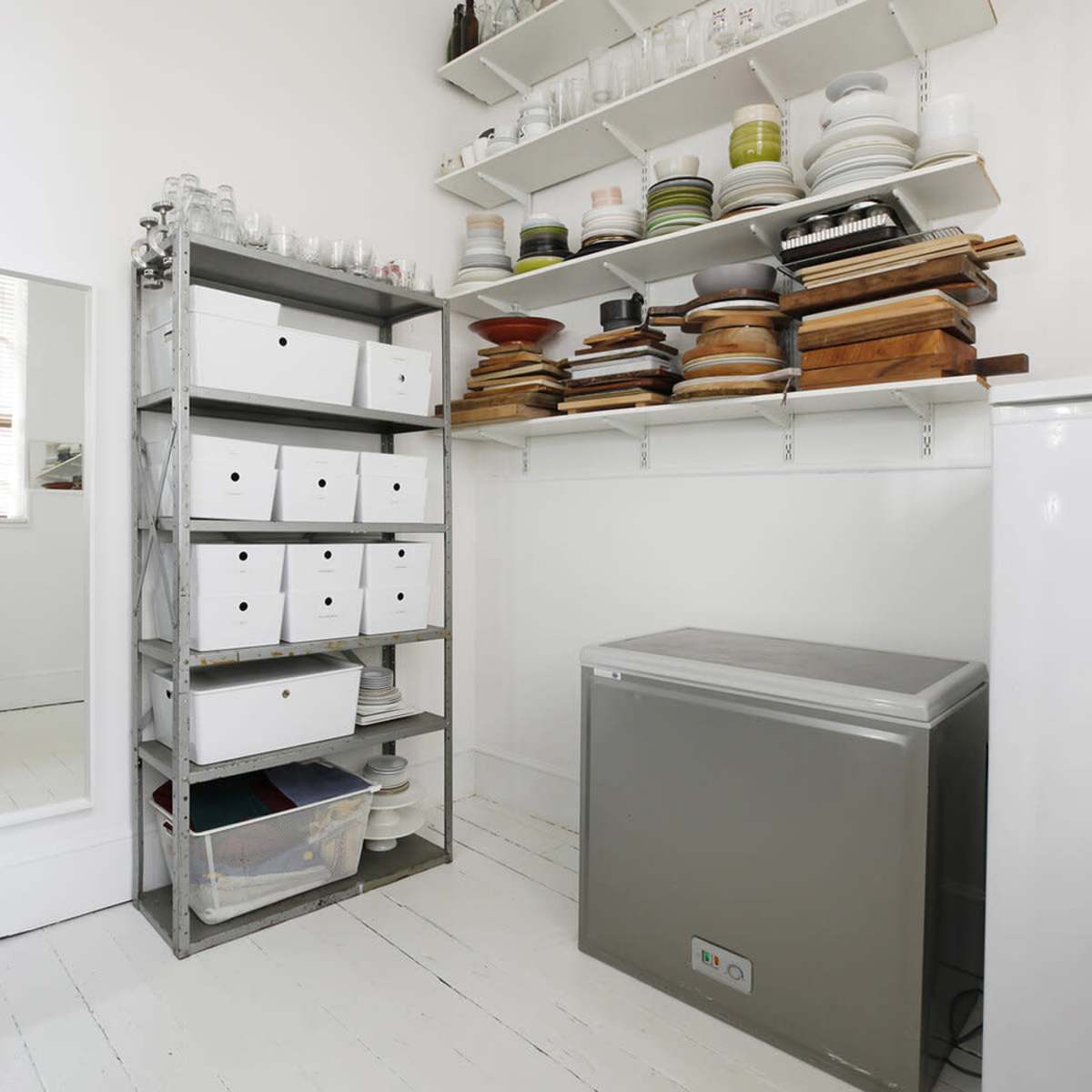 A clean storage area with shelves holding dishes and wooden boards, alongside a metal shelving unit filled with boxes and a chest freezer.