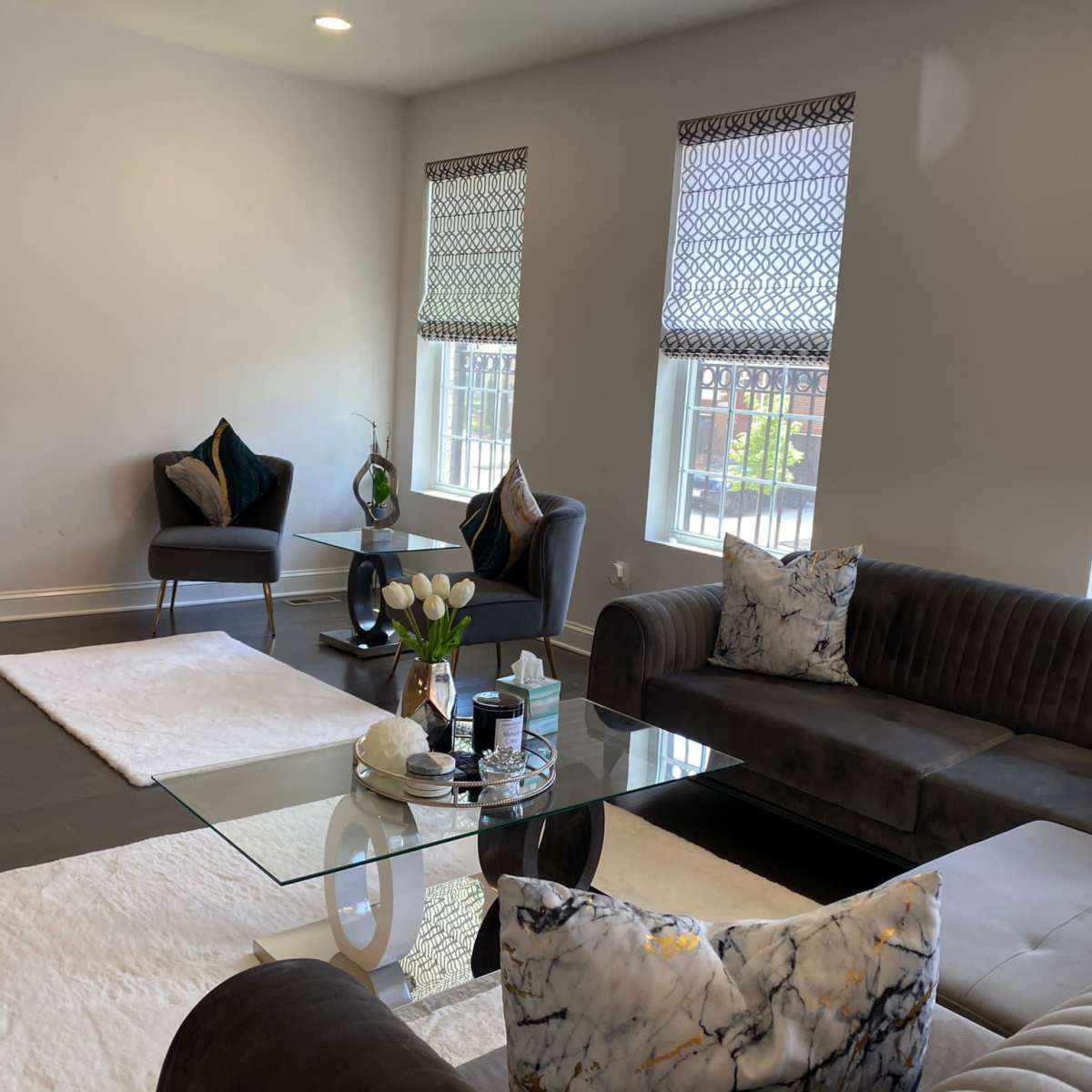 A modern living room with a glass coffee table, dark sofas, and large windows dressed with patterned shades.