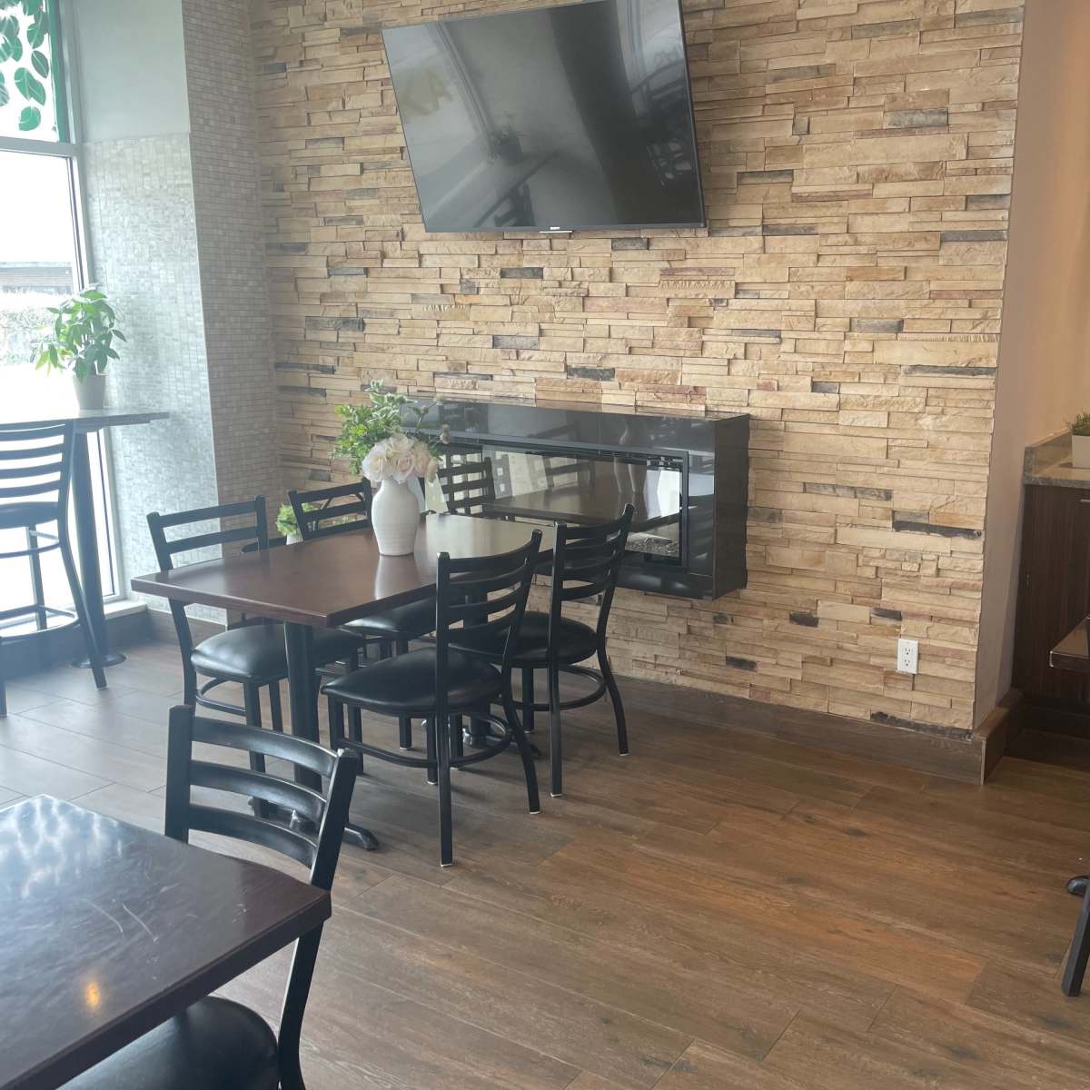 A modern restaurant interior with wooden floors, stone feature walls, black chairs, and a mounted television.
