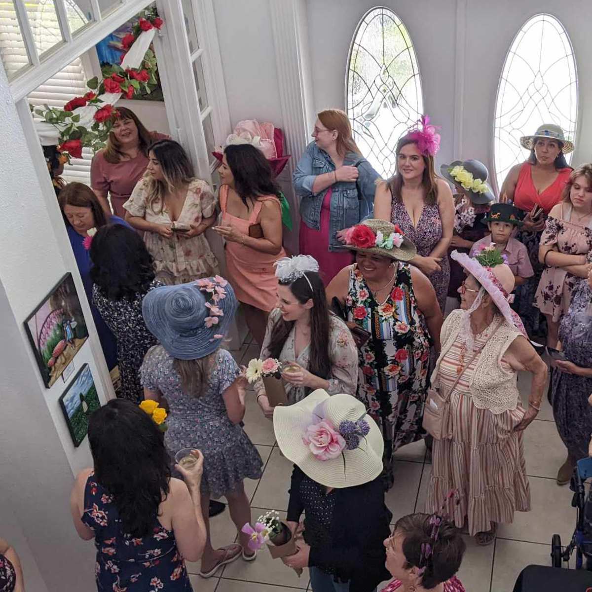 A group of women, many wearing decorated hats, gather in a brightly lit foyer adorned with flowers and photographs.