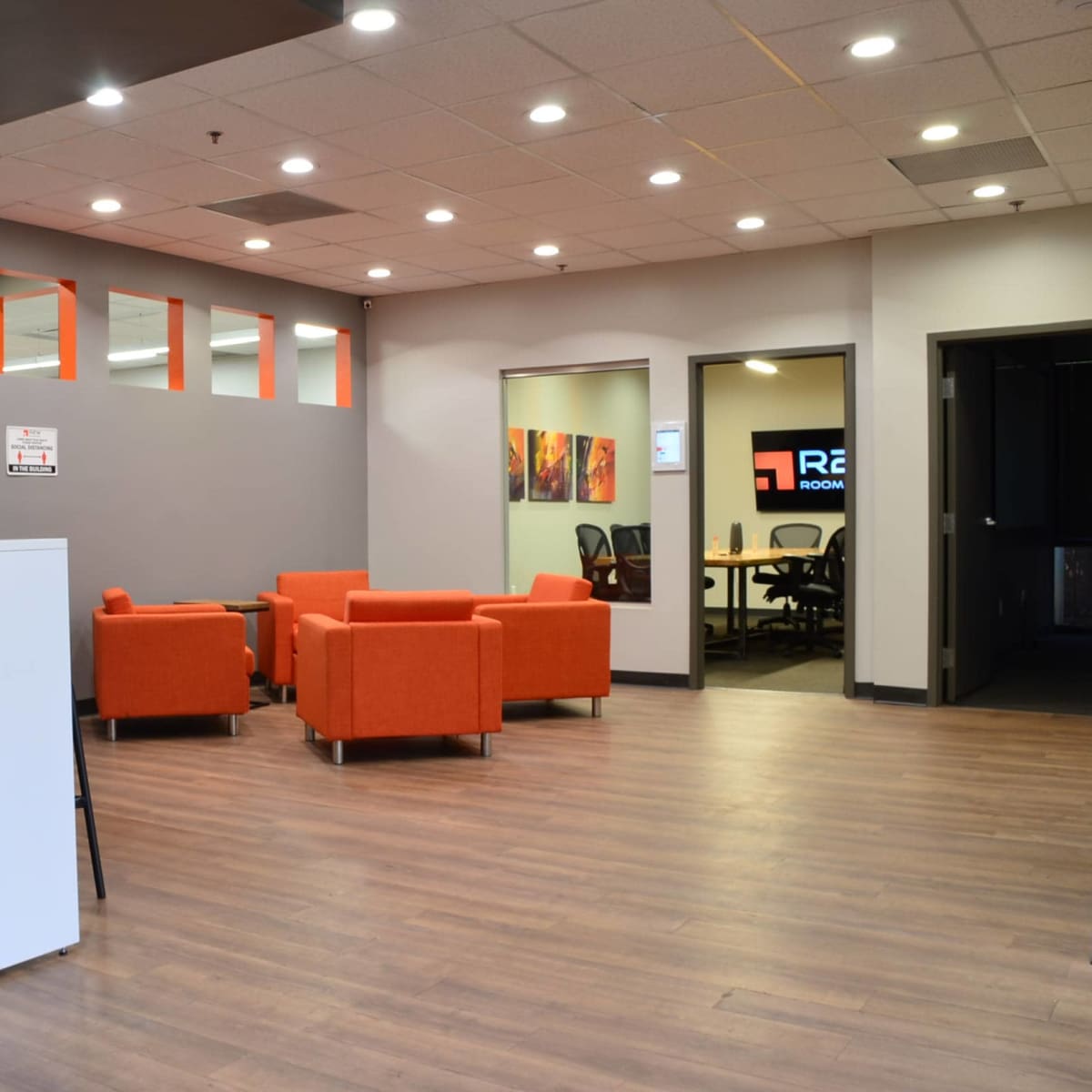 A modern interior space featuring a bar-style counter, orange chairs arranged in seating areas, and a conference room with a visible logo on the wall.