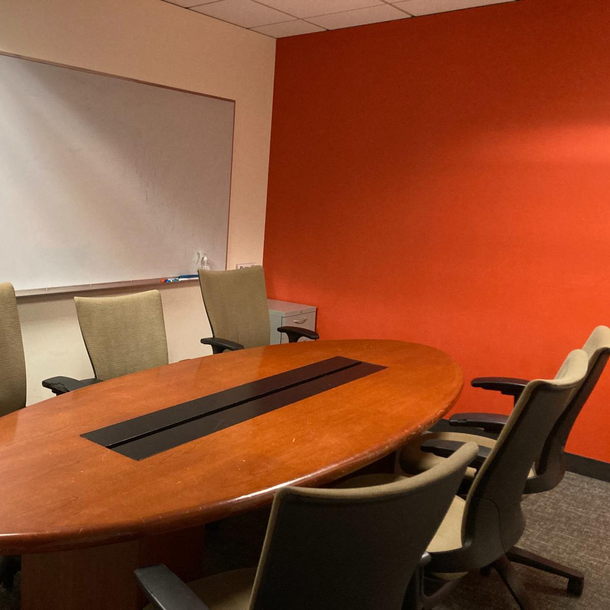 A conference room features a large oval wooden table surrounded by six chairs, with a whiteboard on one wall and an orange accent wall.