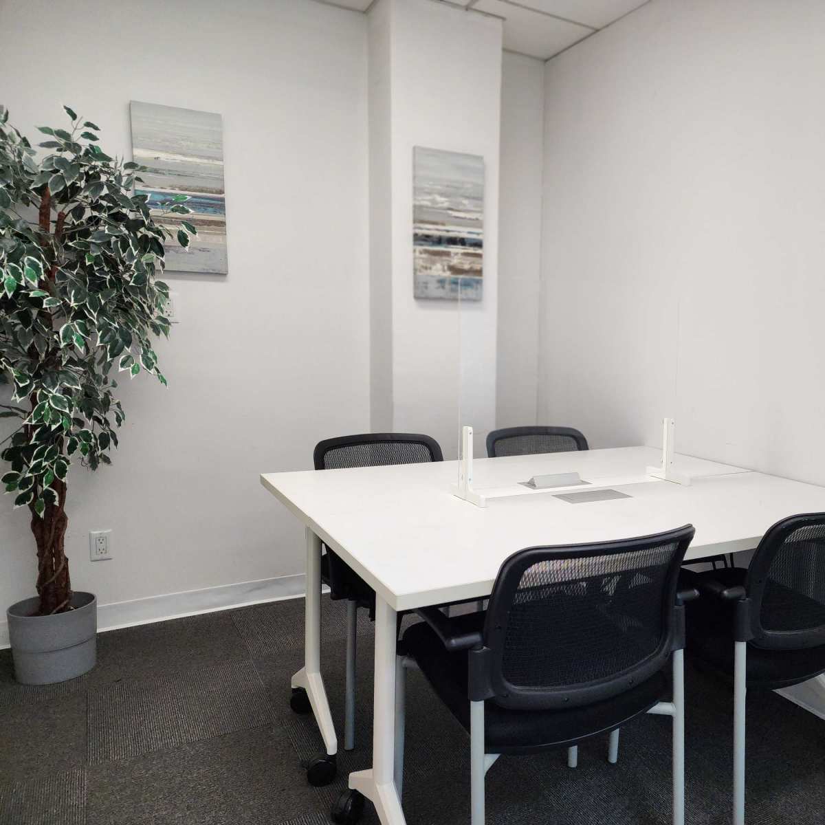 A small, minimalist meeting room featuring a white table surrounded by four black chairs, a potted plant, and two framed artworks on the wall.