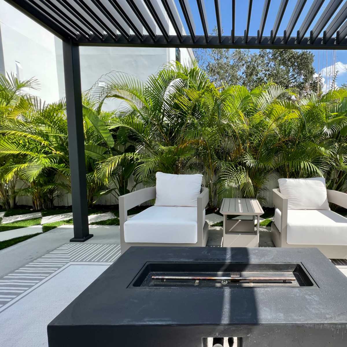 A cozy outdoor seating area features two white chairs and a small table under a pergola, surrounded by lush greenery.