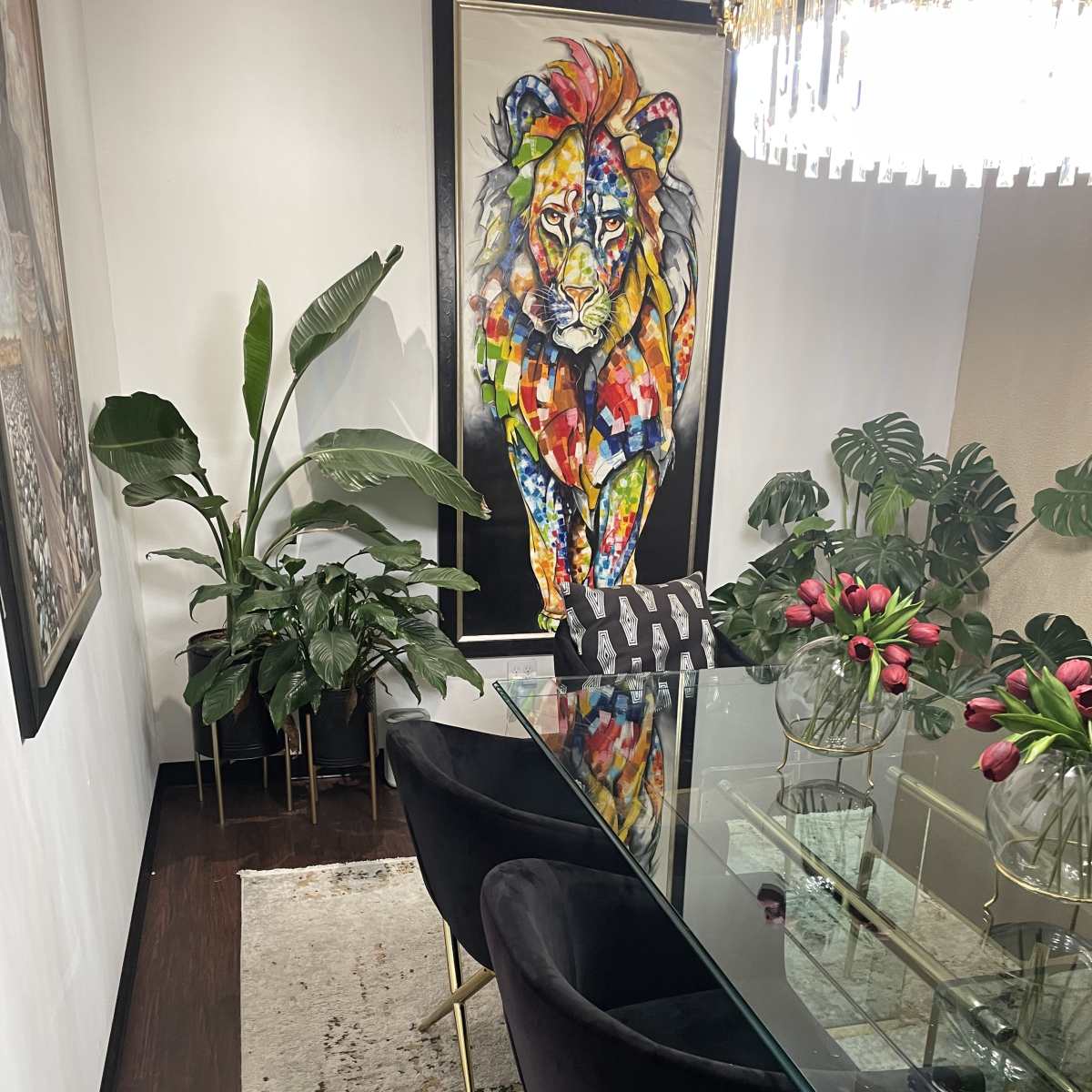 A modern dining area features a glass table surrounded by black chairs, with a colorful lion painting on the wall and lush plants in the corners.