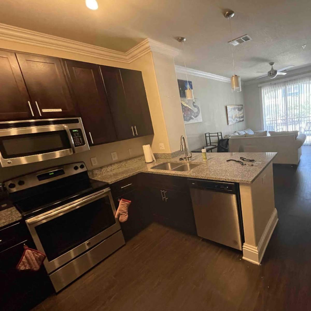 A modern kitchen with dark cabinets and stainless steel appliances, adjacent to a living area with a sofa and large windows.