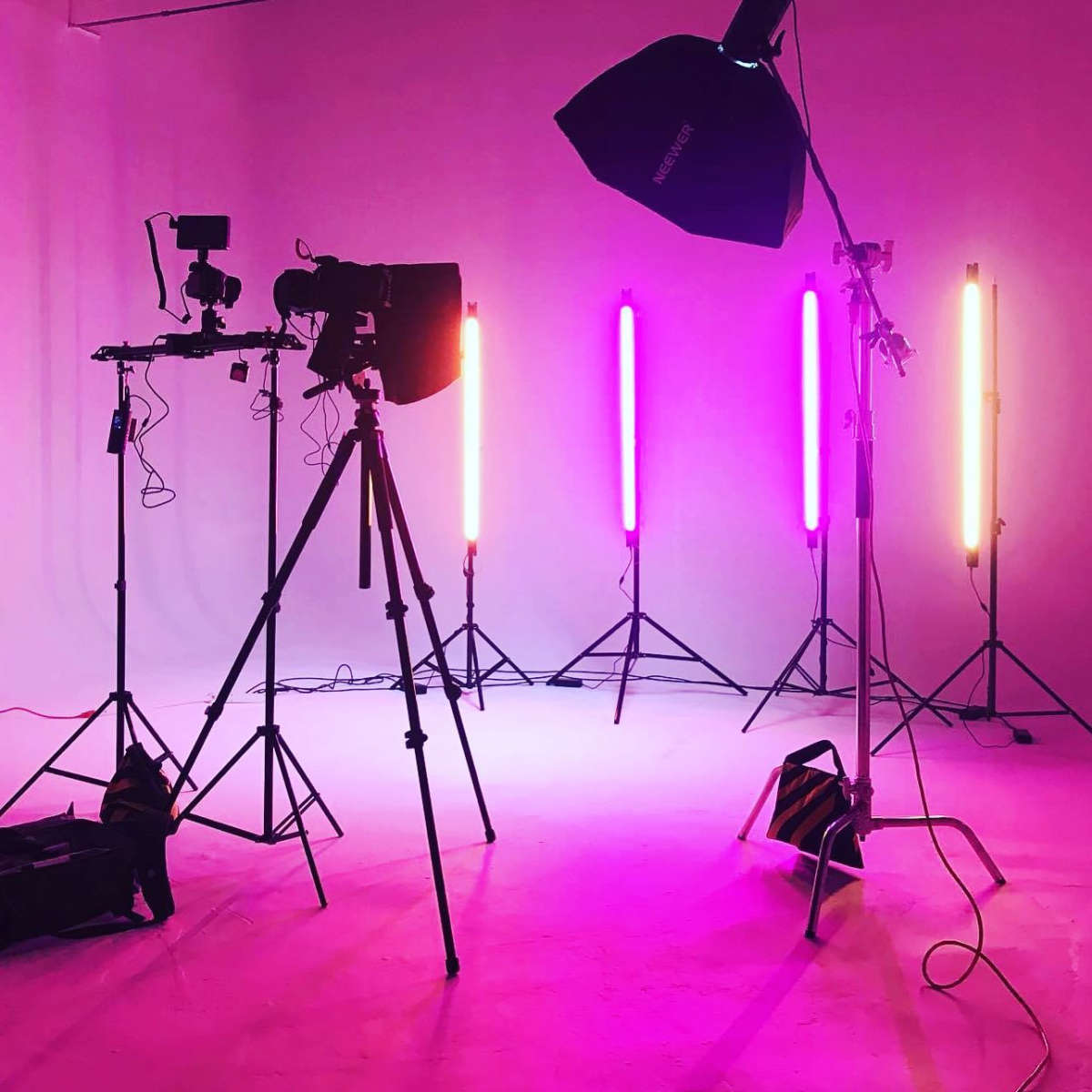 A photography studio setup with multiple tripods, lighting equipment, and colored light tubes against a pink backdrop.