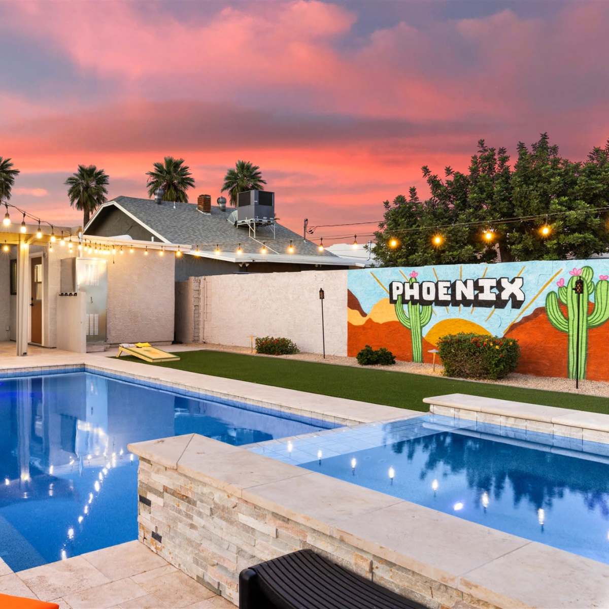 A swimming pool with lounge chairs is illuminated by string lights against a backdrop of a sunset sky and a mural reading "PHOENIX."