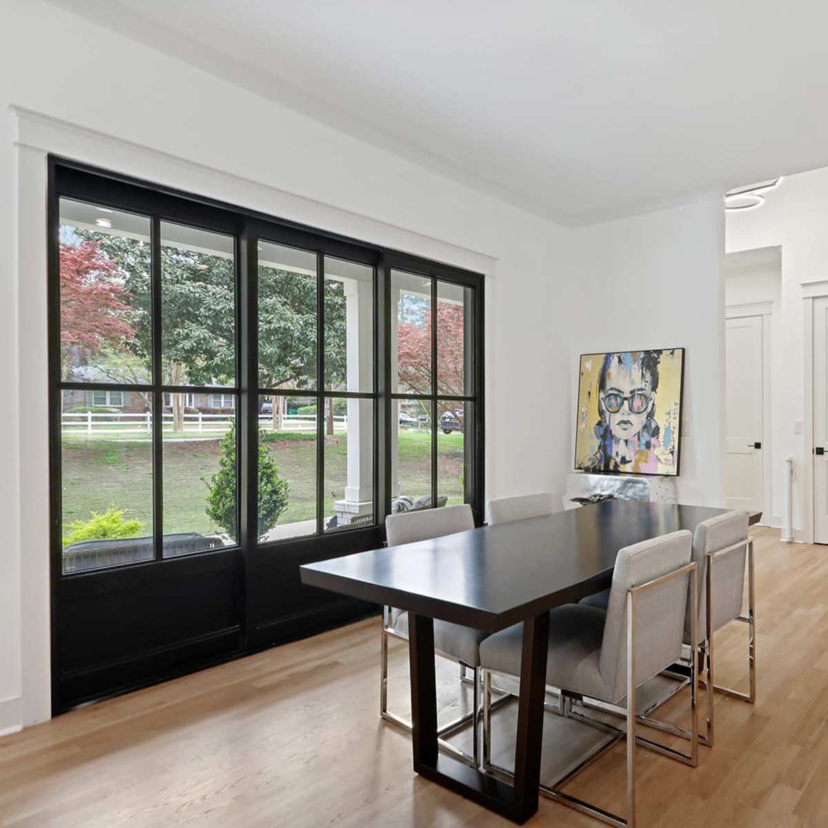A dining area features a black table surrounded by silver chairs, with large windows overlooking a landscaped yard.
