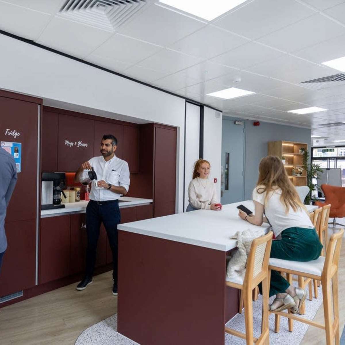 A modern office break room features a coffee station with several people engaging in conversation and using their devices.