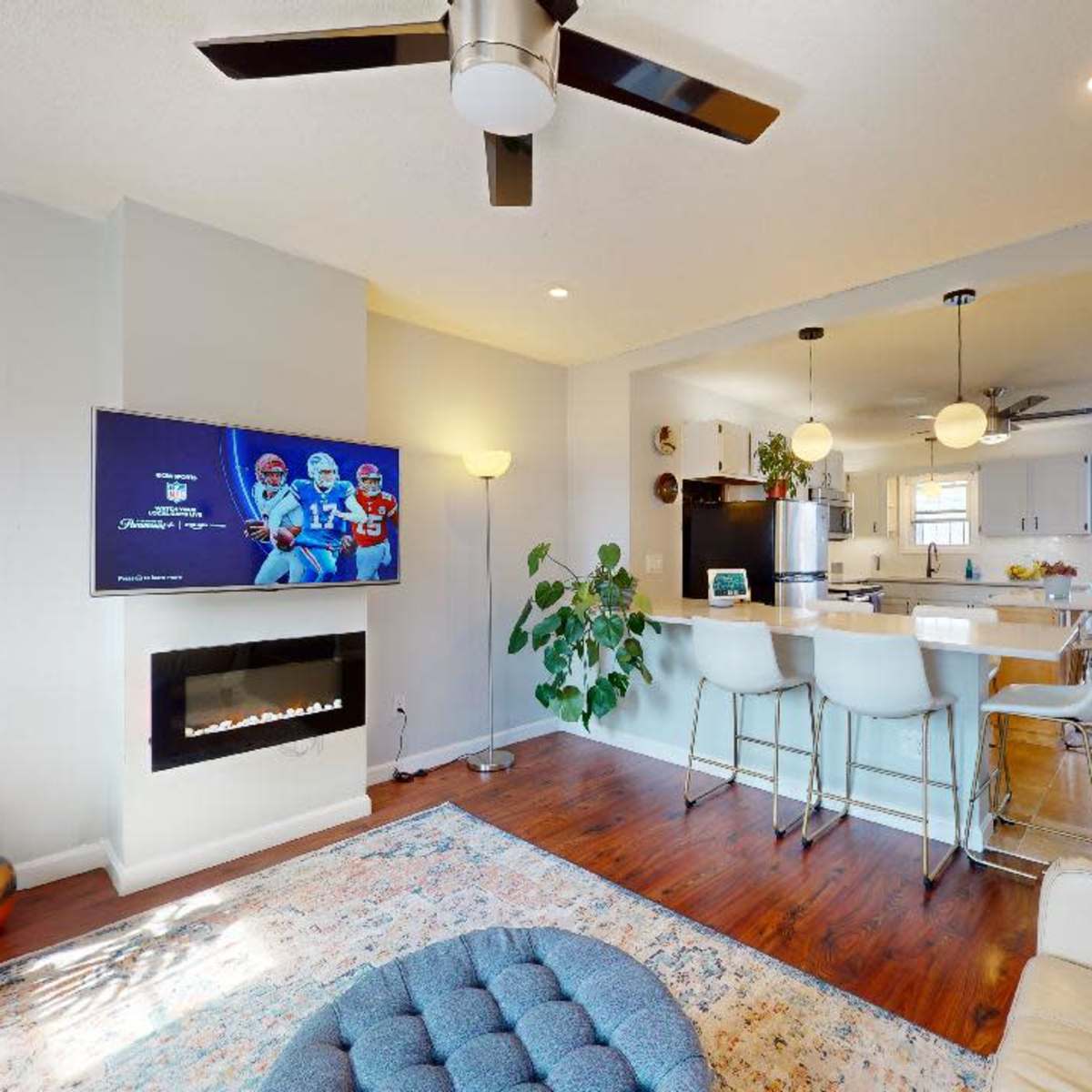 A modern living space with a wall-mounted television, a ceiling fan, a kitchen bar with stools, and a fireplace surrounded by light-colored walls and wooden flooring.