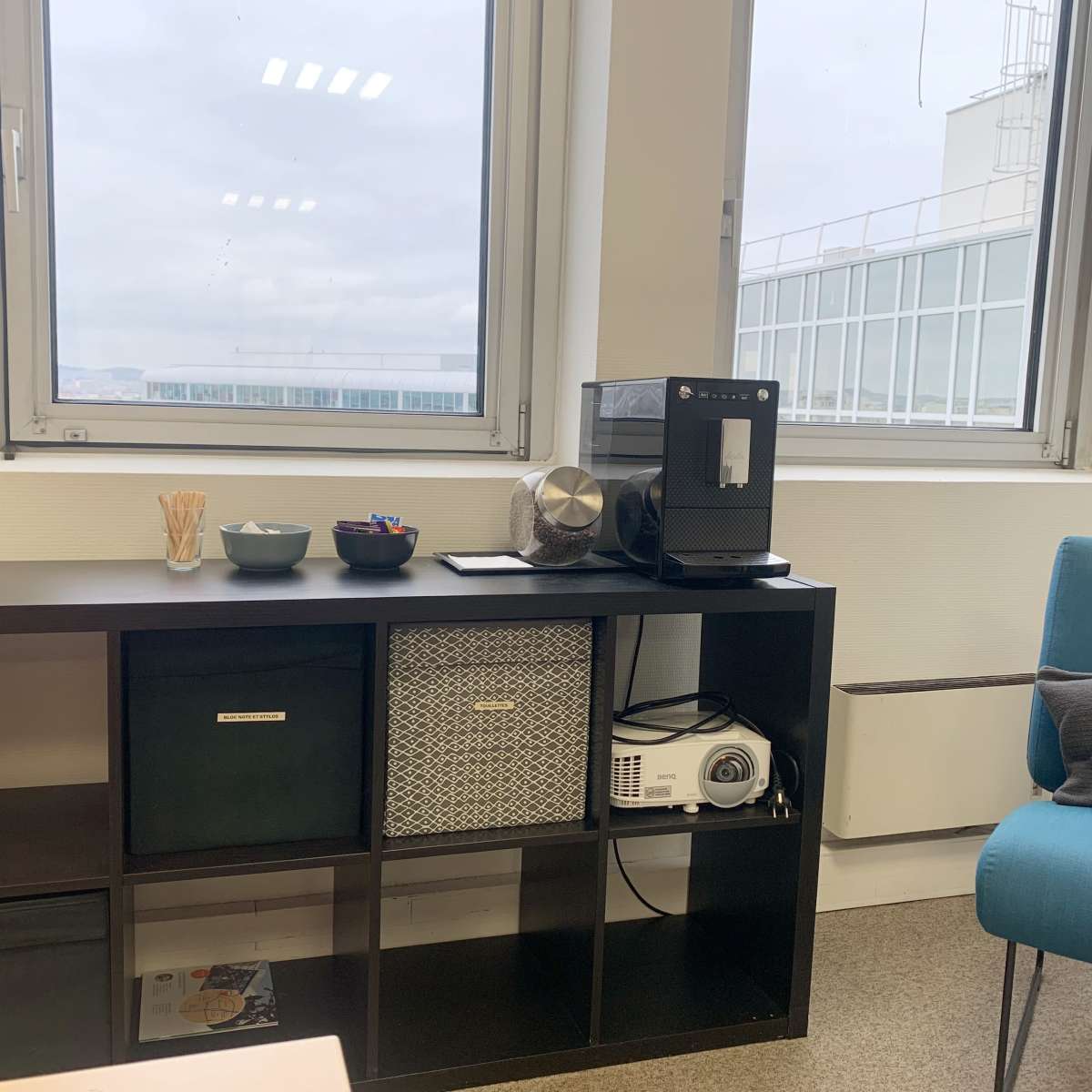 A workspace with a shelf holding a coffee machine, a projector, and storage boxes, alongside a blue chair and large windows overlooking an outdoor view.