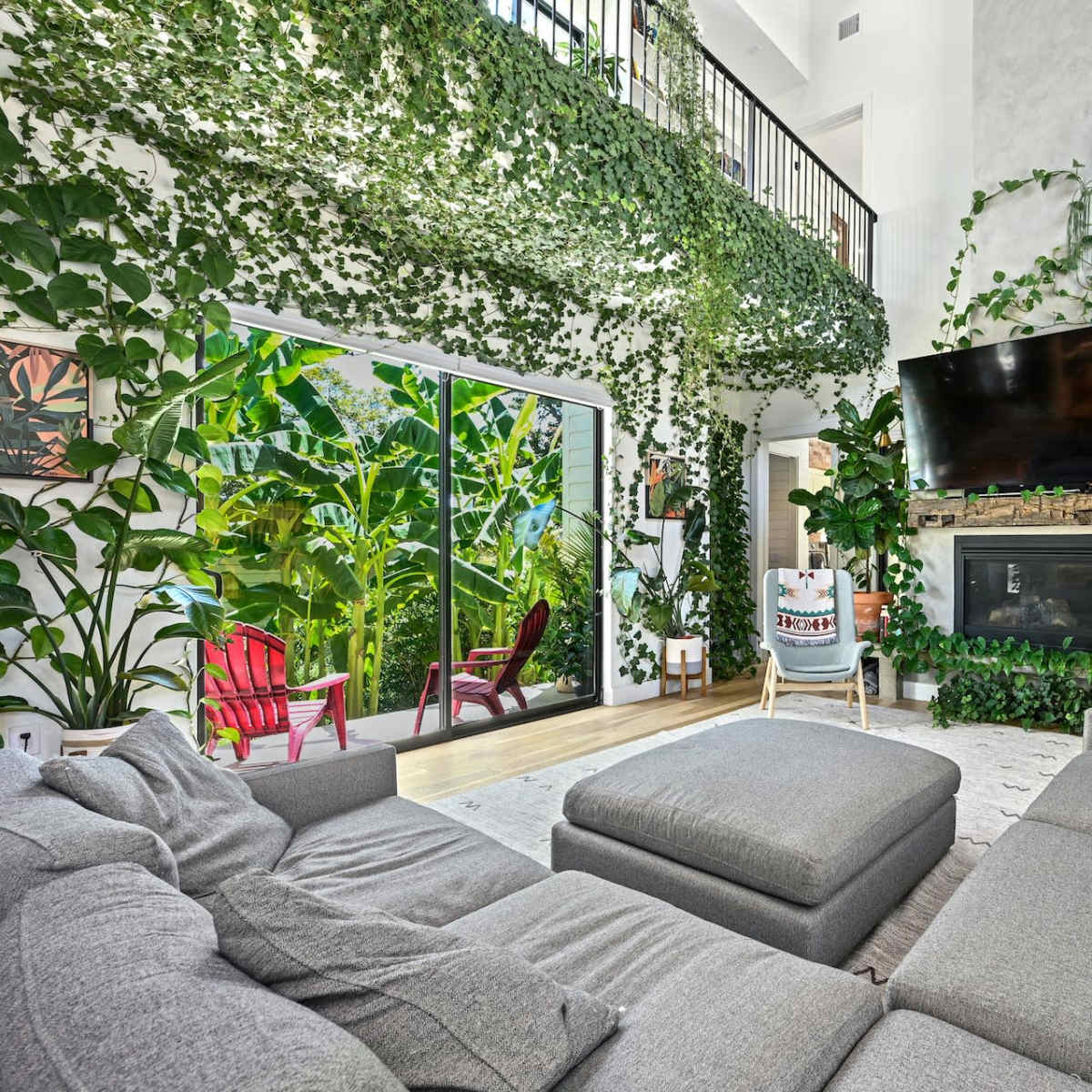 The living room features a large sectional sofa, a mounted TV above a fireplace, and abundant indoor plants surrounding glass doors that open to a lush outdoor environment.