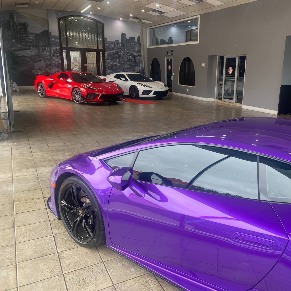 A car showroom featuring a purple Lamborghini in the foreground and two other sports cars, one red and one white, in the background.