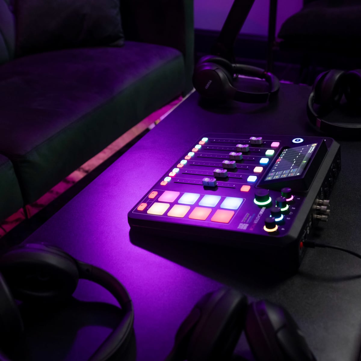 A music production controller and several pairs of headphones rest on a black table beside a green couch in a dimly lit room.