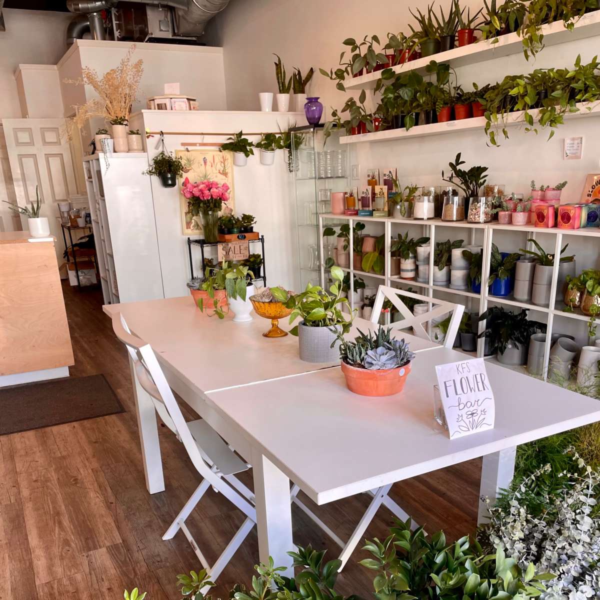 A bright, organized shop features white tables adorned with plants and floral arrangements, surrounded by shelves filled with various greenery and decorative items.