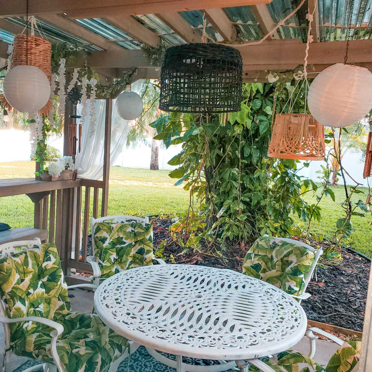 A gazebo with a round white metal table surrounded by patterned chairs, adorned with hanging lanterns and greenery.