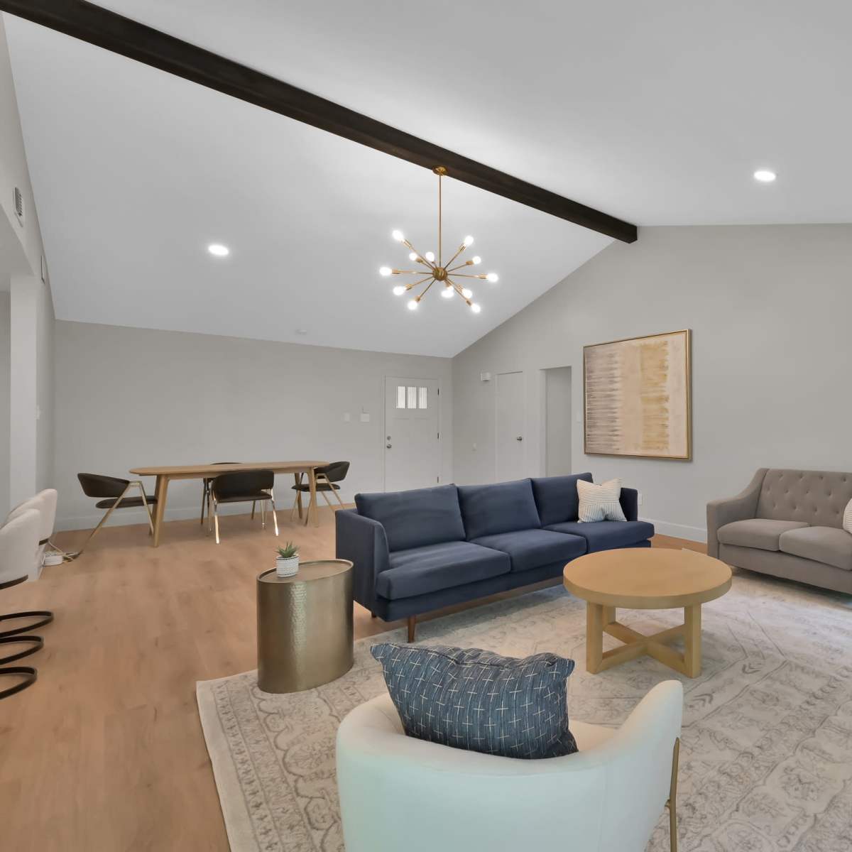 A modern living room featuring a blue sofa, a light gray couch, a round wooden coffee table, and a decorative plant, with a dining area visible in the background.