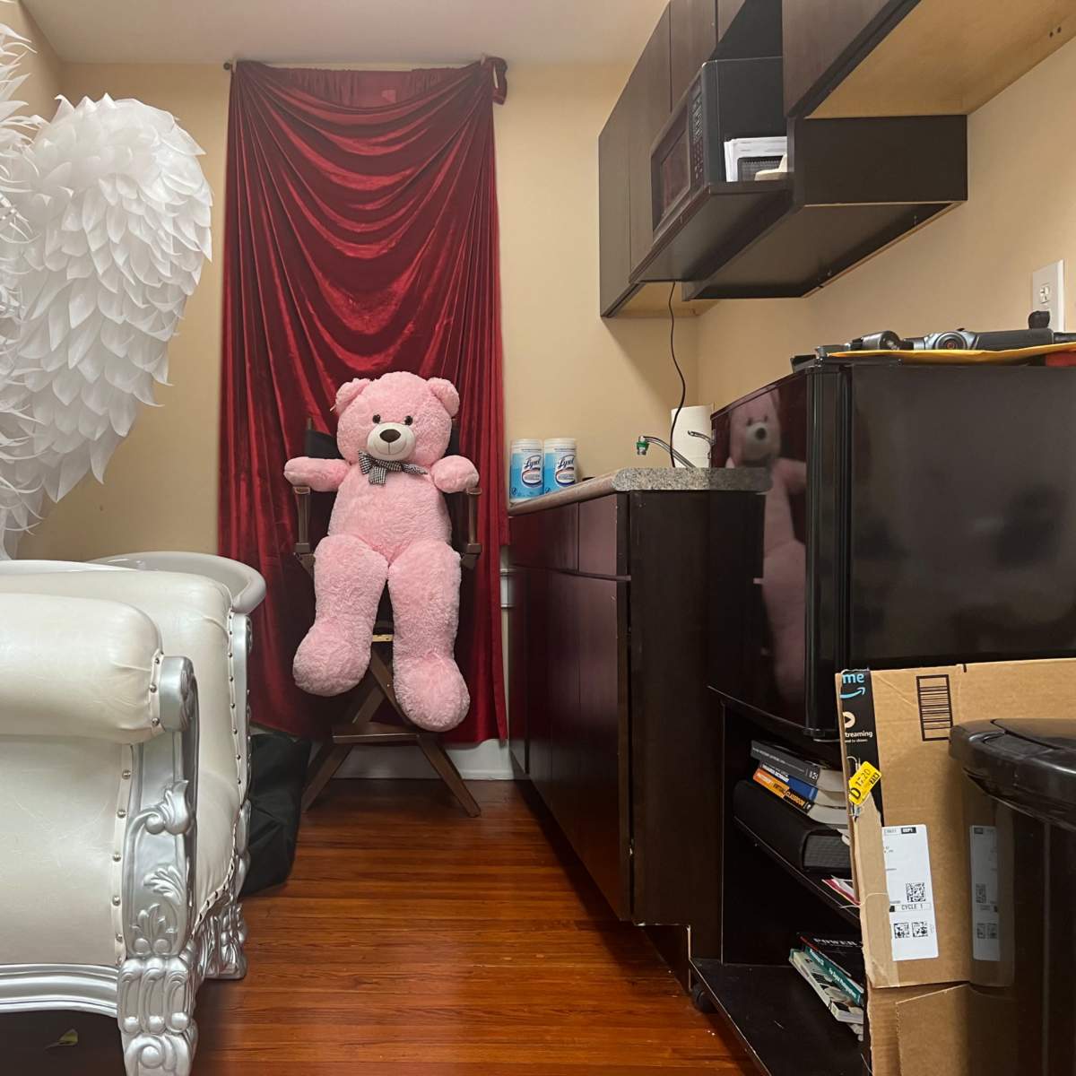 A small room with a large white chair, a pink teddy bear sitting on a stool, and a red curtain in the background.