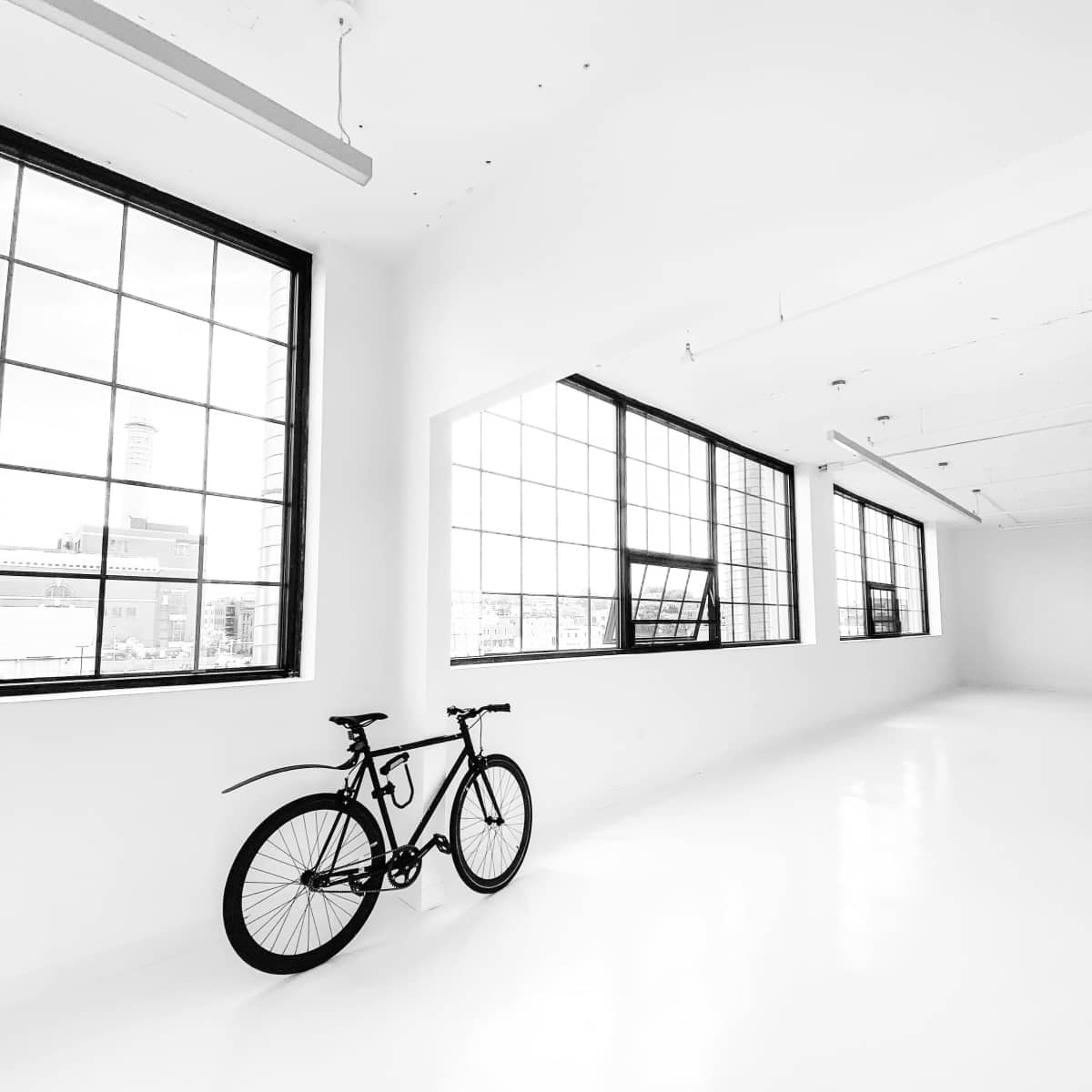 A black bicycle leans against a white wall in a spacious, well-lit room with large windows.