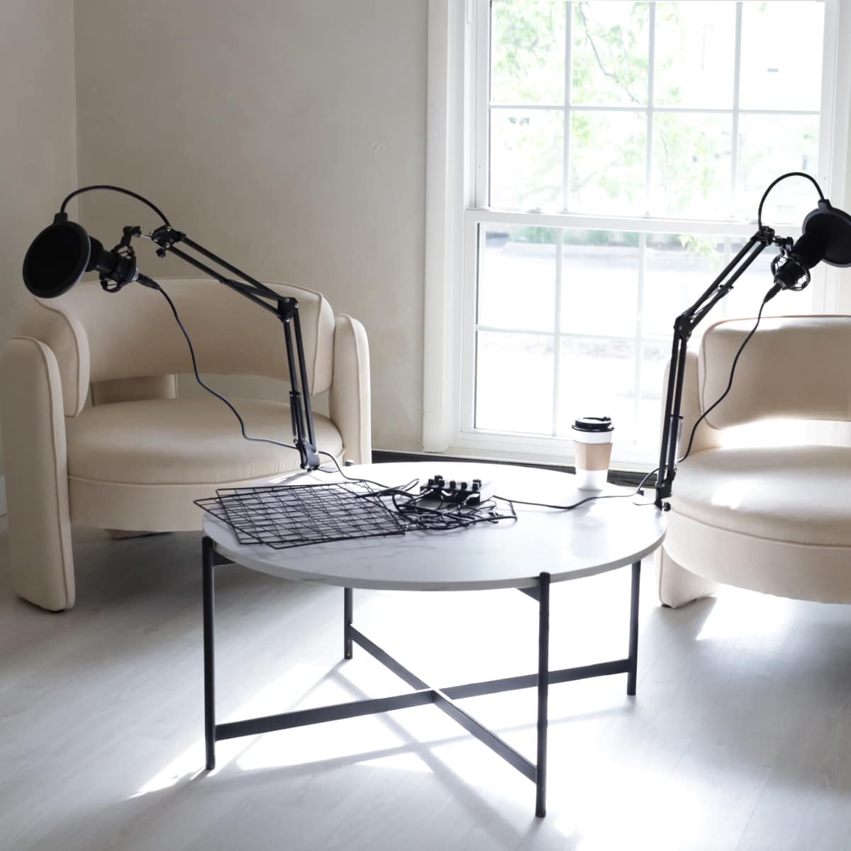 Two cream-colored chairs with adjustable microphone stands beside a round white coffee table in a well-lit room with a large window.