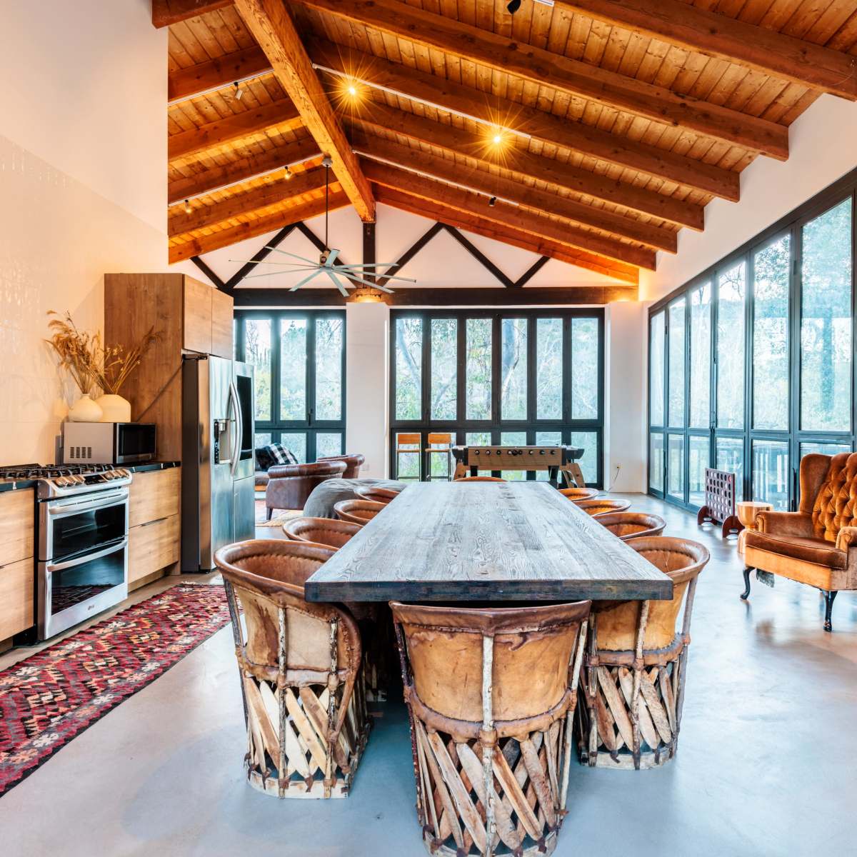 A spacious dining room features a large wooden table surrounded by woven chairs, with a high ceiling supported by exposed beams and large windows allowing for ample natural light.