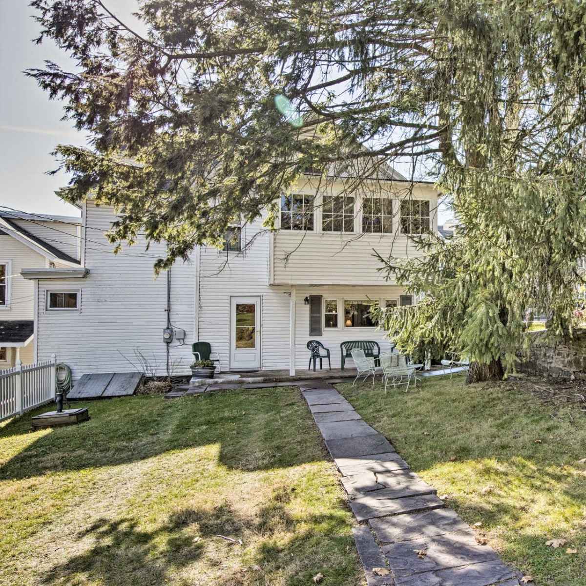 A two-story white house with a backyard featuring a path made of stones, a grassy area, and a large tree.