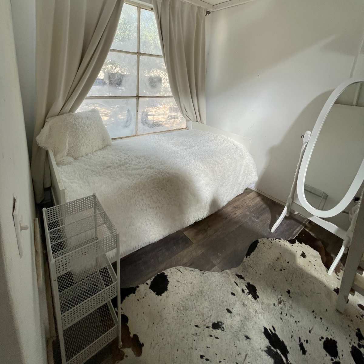 A small bedroom with a single bed, a white mirror, a wire storage unit, and a window with light curtains, set on wooden flooring with a cowhide rug.
