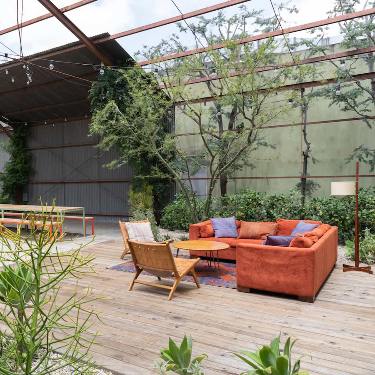A modern outdoor seating area features a rusted metal framework surrounded by greenery, with a couch, two chairs, and a long table.
