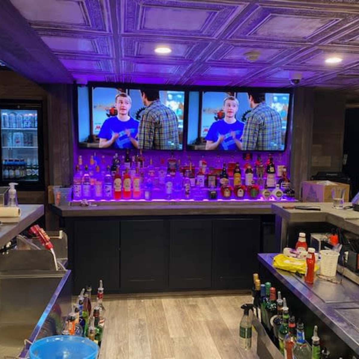 A bar area with a well-stocked liquor shelf, two televisions displaying a program, and various condiments and supplies on the counter.
