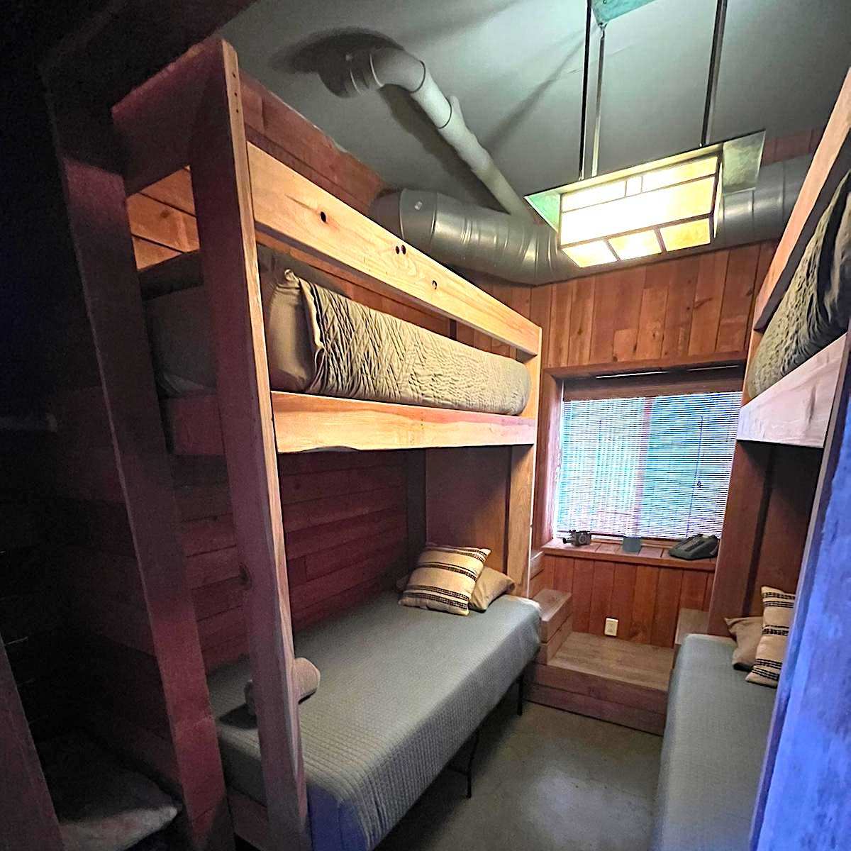 A small cabin room featuring two sets of wooden bunk beds and a window with a green light fixture overhead.