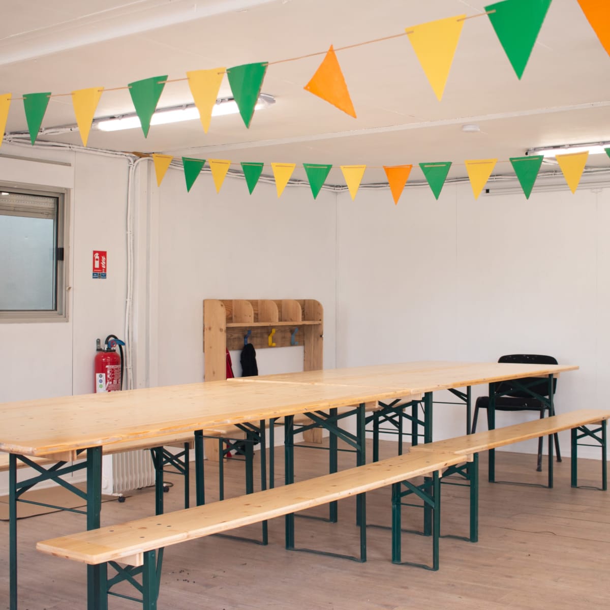 A spacious room features a long wooden table with benches and decorative green and orange bunting overhead.