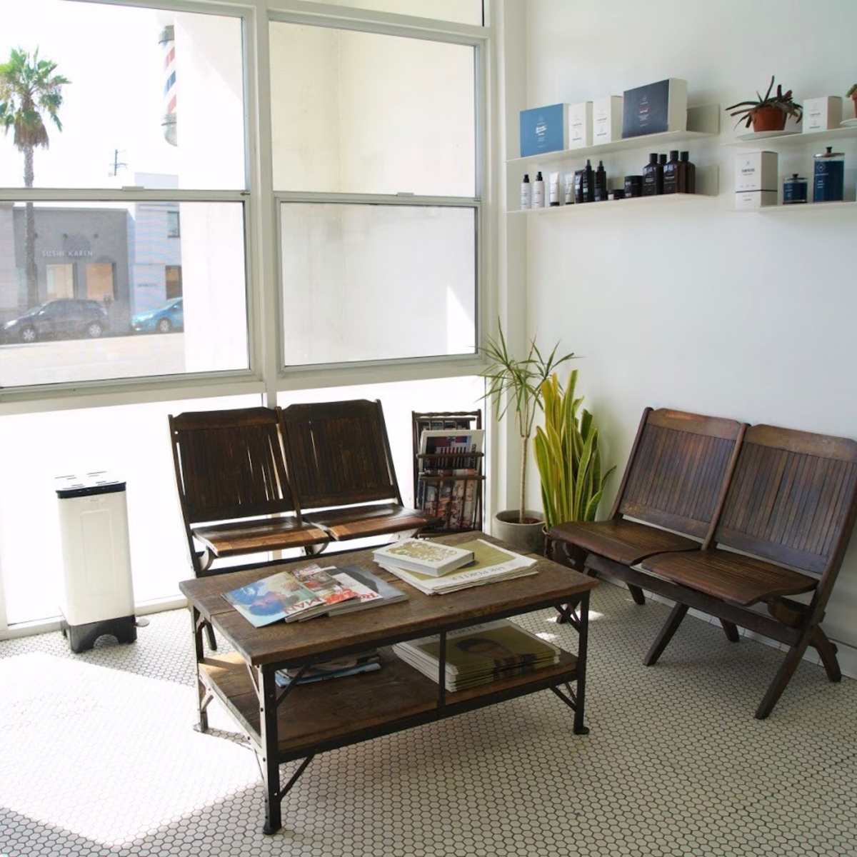 The interior of a barbershop features two wooden chairs and a coffee table, with large windows allowing natural light to illuminate the space.