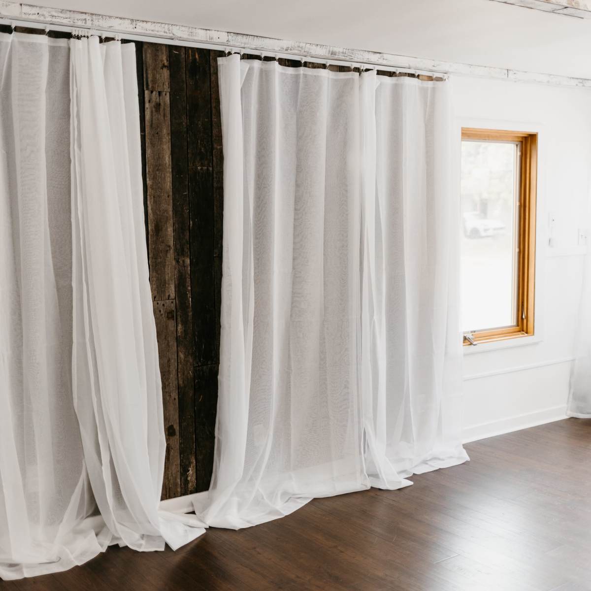 Sheer white curtains drape elegantly over a wooden beam, framing a window in a bright, minimalist room with wooden flooring.